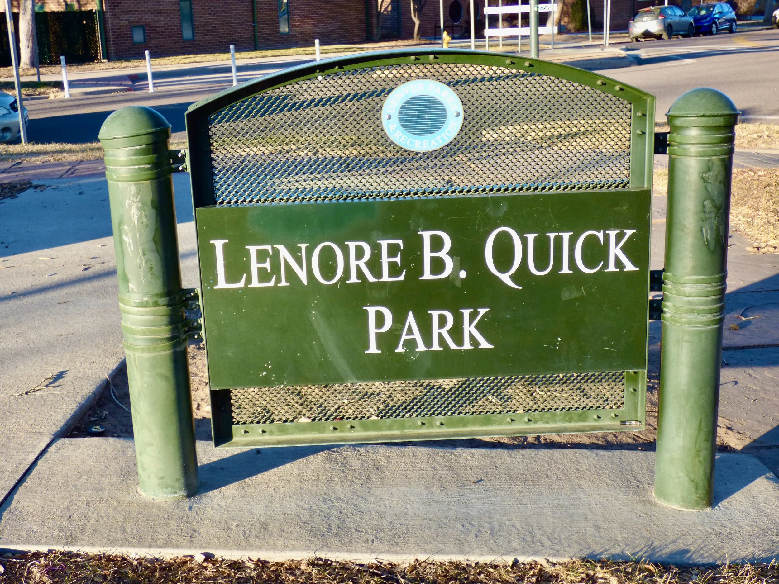 Sign for Lenore B. Quick Park in a city setting, with cars parked in the background and a sidewalk in the foreground.