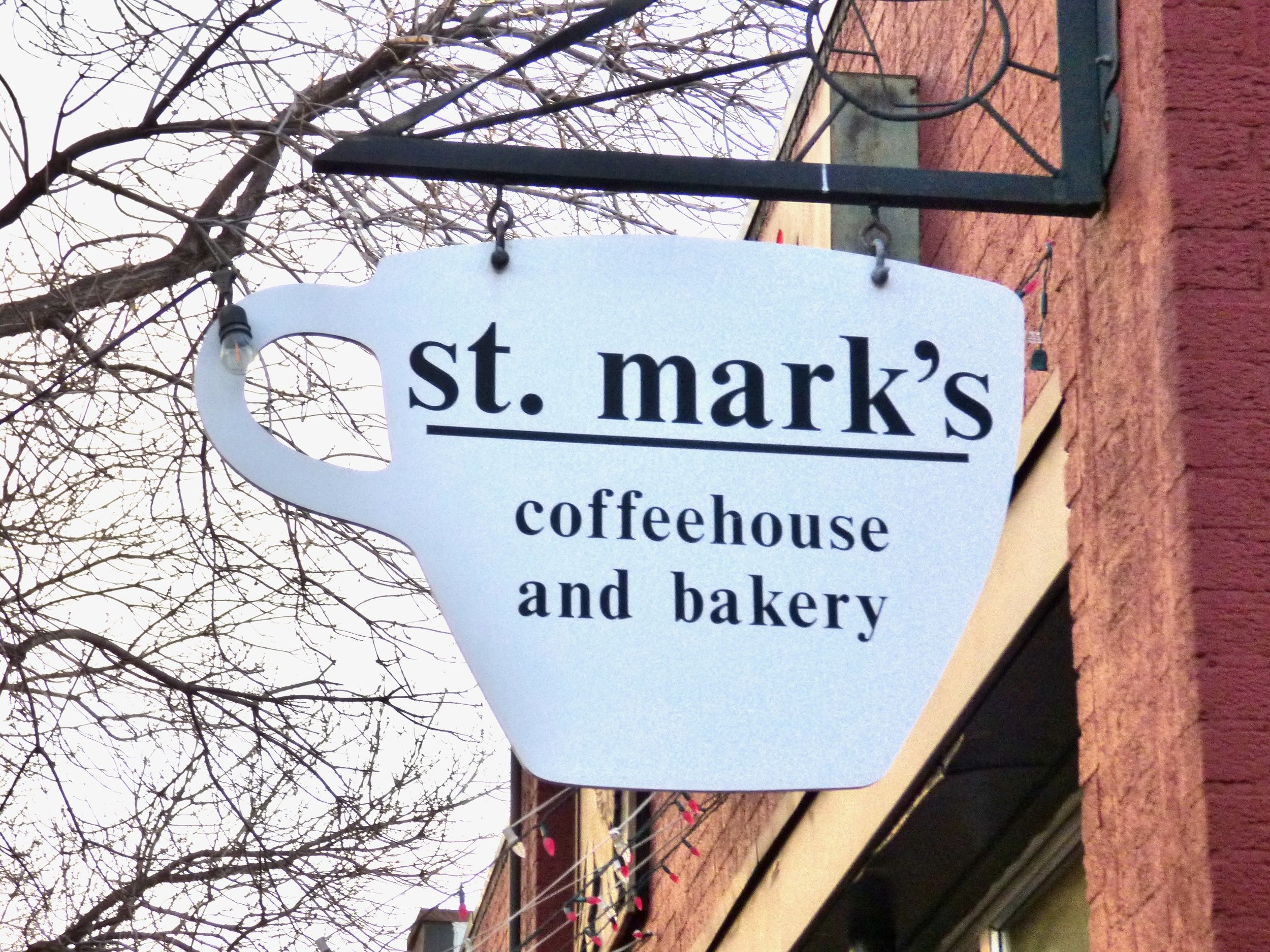 A white coffee mug-shaped sign reads 'st. mark's coffeehouse and bakery,' hanging on a black metal bracket outside a brick building, with leafless tree branches in the background at St. Mark's Coffeehouse and Bakery, Denver, CO.