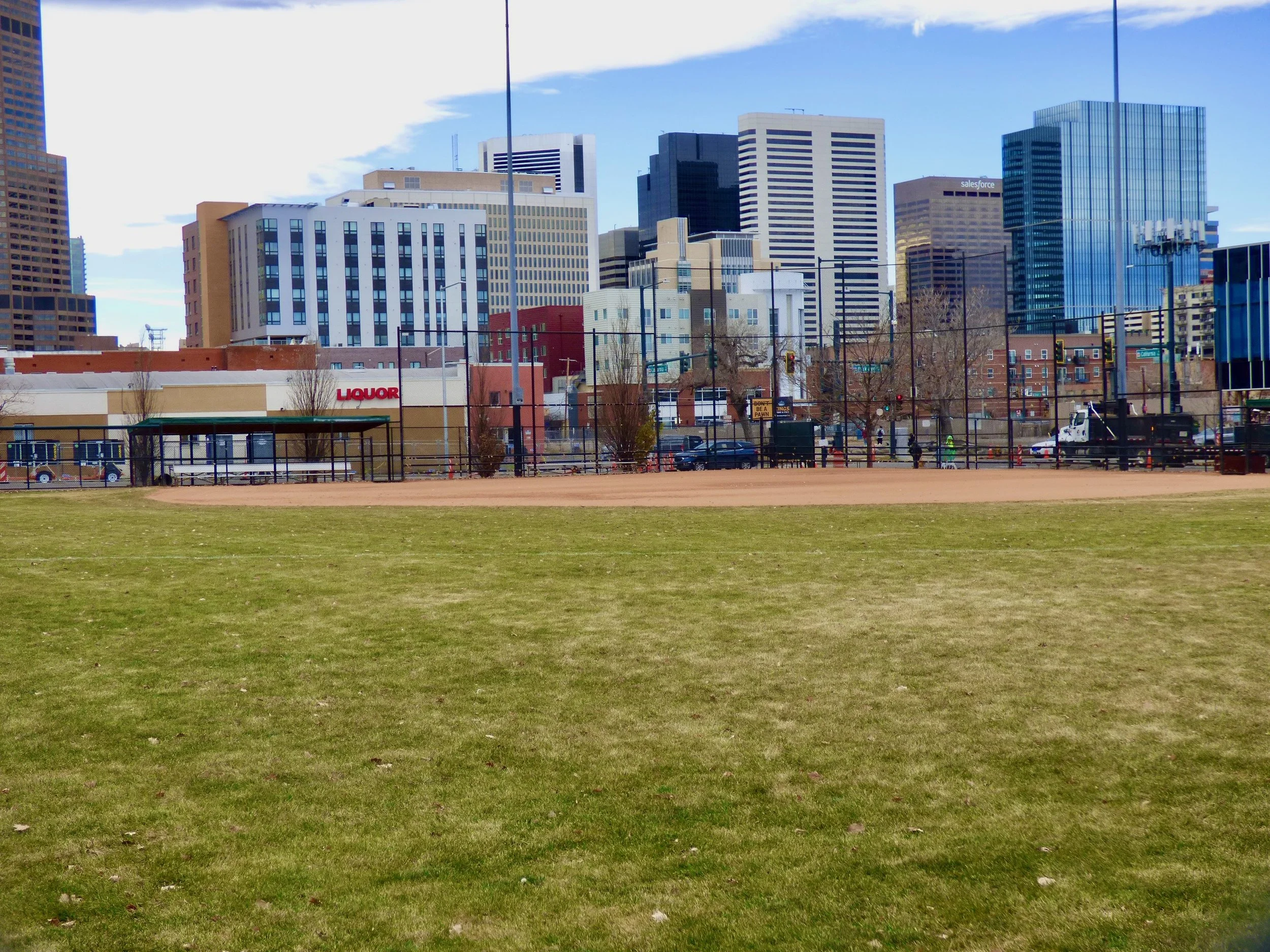A cityscape with tall modern buildings in the background and a baseball field with green grass and a dirt infield in the foreground. There are some trees, people, and parked cars near the buildings in Sonny Lawson Park, Denver, CO 80205.