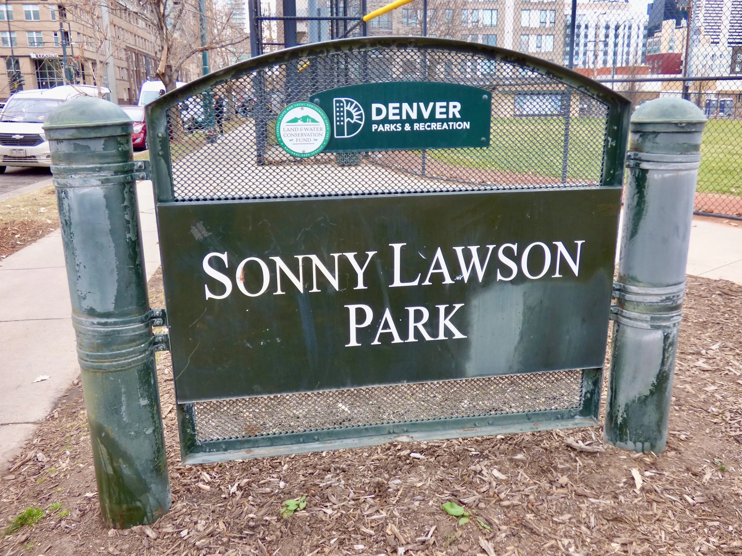 Park sign for Sonny Lawson Park in Denver, Colorado, with city buildings and park fencing in the background in Sonny Lawson Park, Denver, CO 80205.