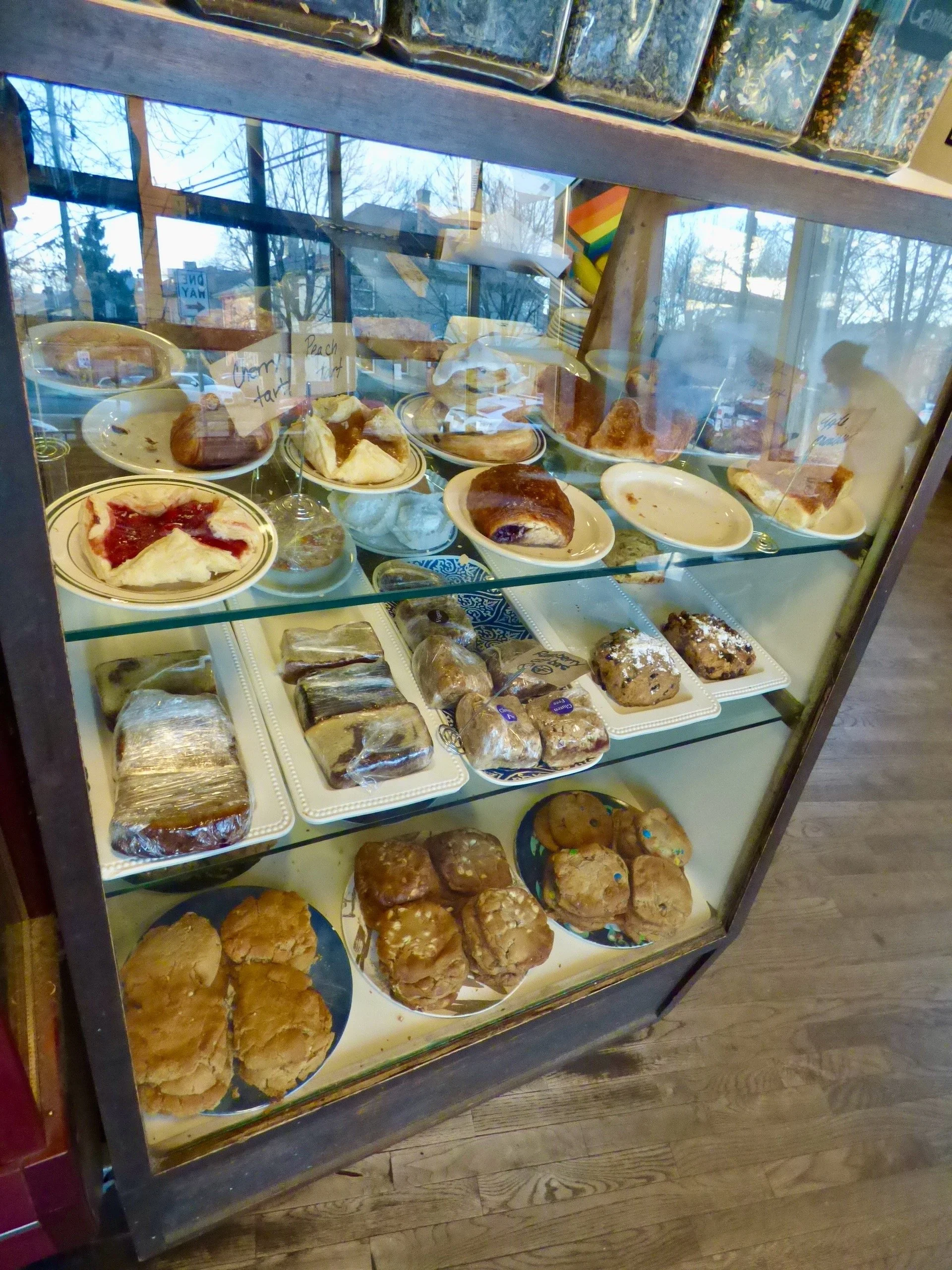 Display case filled with various baked goods including pies, scones, cookies, and pastries, with handwritten signs inside the case at St. Mark's Coffeehouse and Bakery, Denver, CO.