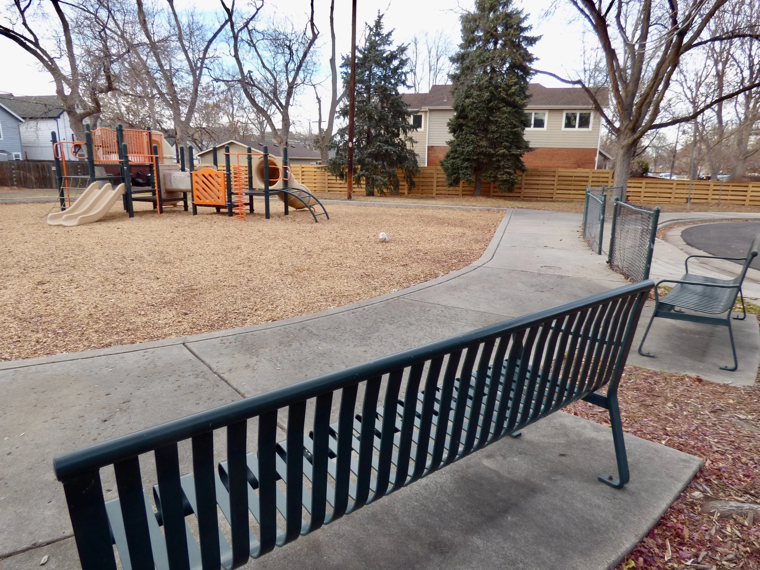Empty playground with a playground structure, benches, trees, and a surrounding fence at Dr. Daniel Hale Williams Park, Denver, CO 80205.