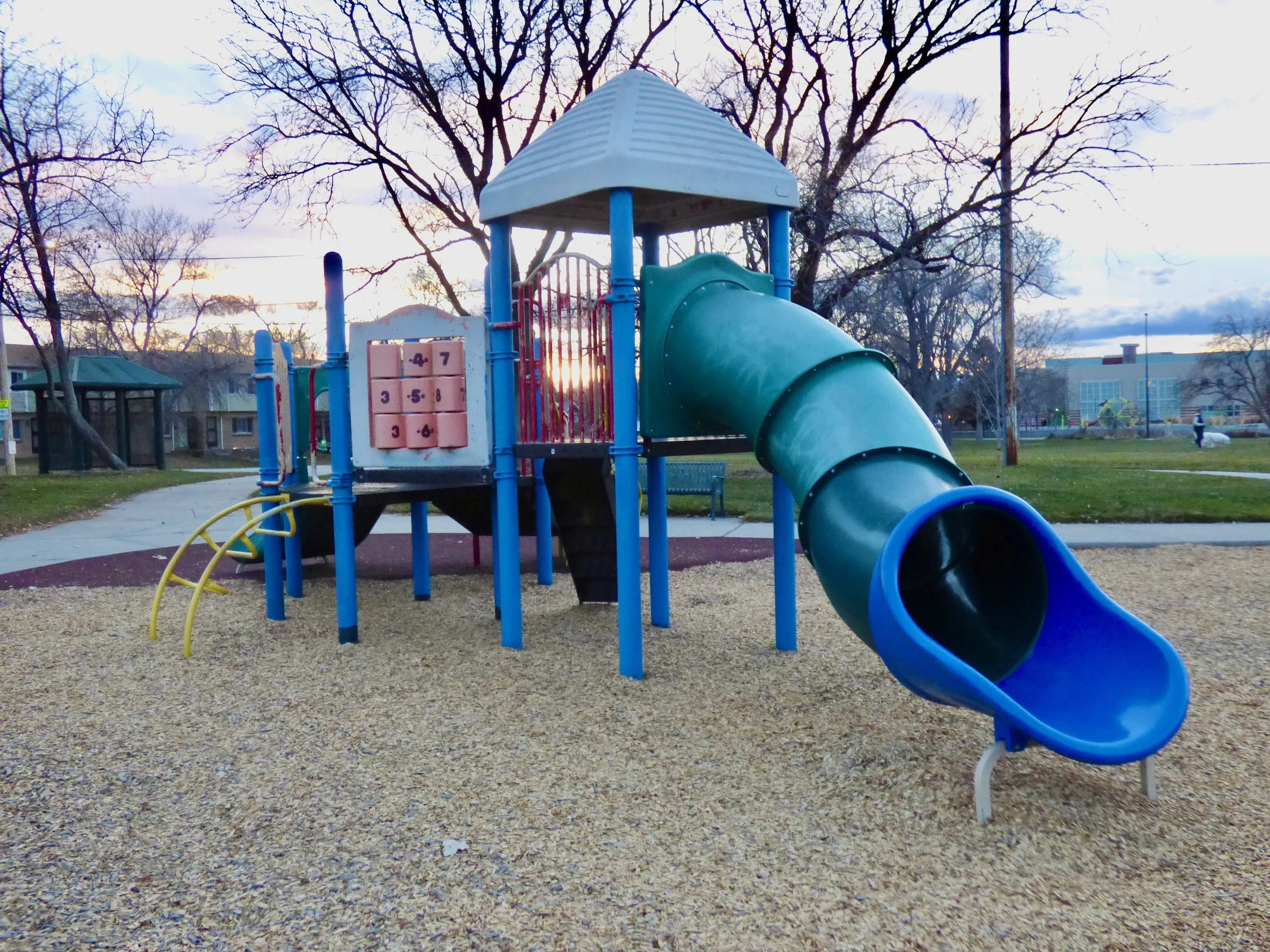 Empty children's playground with a slide, climbing features, and tic-tac-toe game, surrounded by grass and trees, during sunset or sunrise in City of Nairobi Park, Denver, CO 80205.