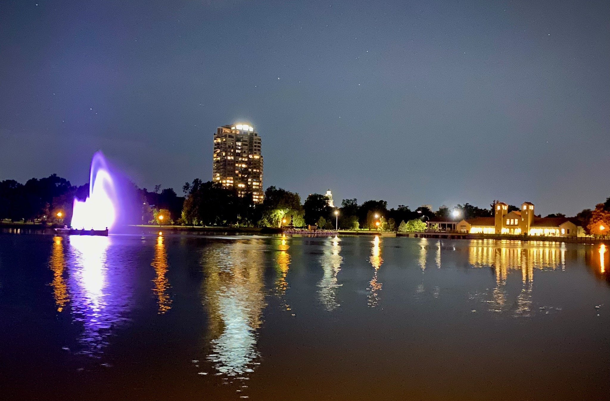 Nighttime cityscape with illuminated fountain on a lake, reflections on the water, tall building with lights, and other buildings with lights in City Park, Denver, CO.