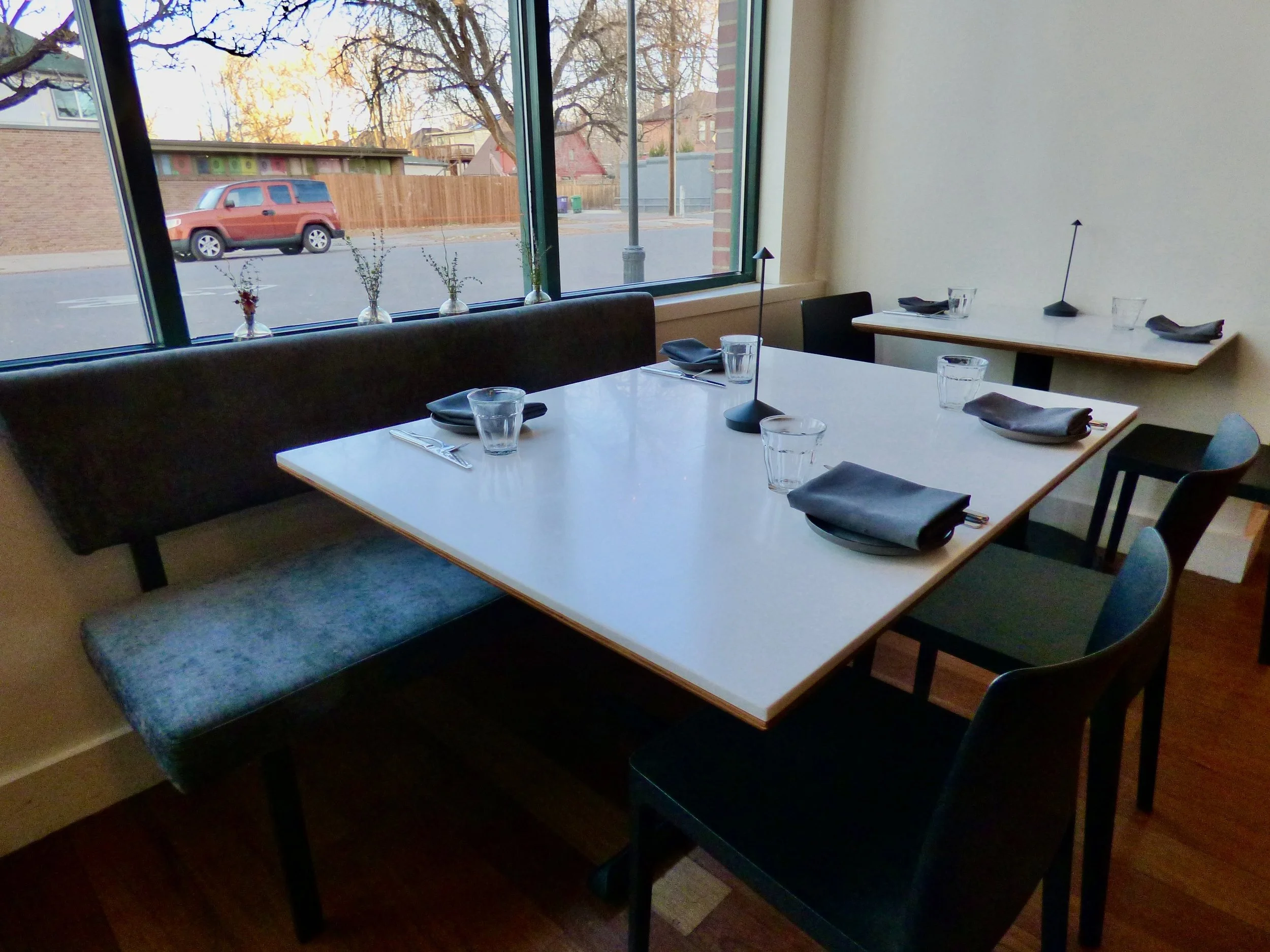 Empty restaurant table set with black napkins, clear drinking glasses, black utensils, black chairs, and a bench in front of large windows, outside view of parked vehicle and trees at Point Easy, Denver, CO 80205.