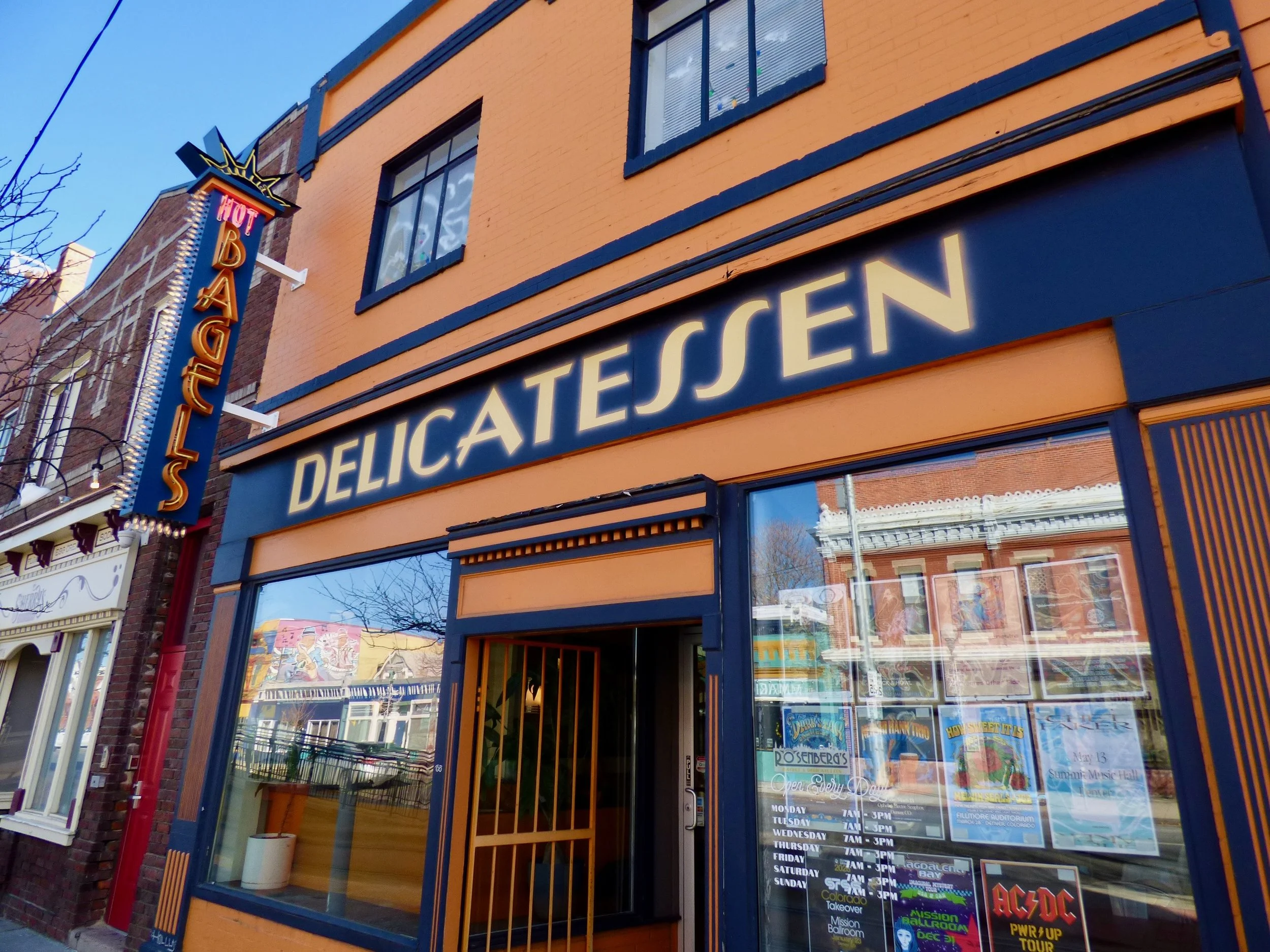 Facade of a delicatessen shop with a large illuminated sign reading 'DELICATESSEN' in yellow letters and a vertical neon sign that says 'HOT BAGELS' on the side of the building.