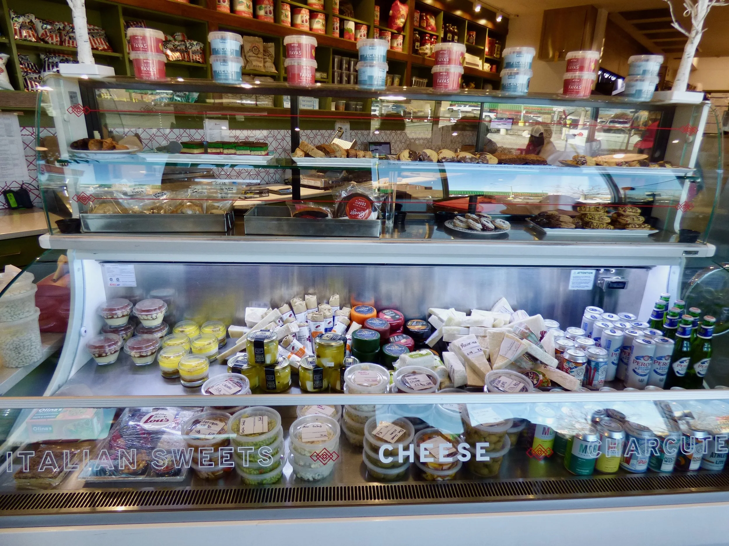 Display case with various cheeses, cheeses, and dairy products, with shelves of packaged sweets and other food items in the background, labeled "Italian Sweets," "Cheese," and "M. Marcute."