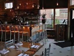 Interior of a restaurant with a bar and tables set for dining, large windows, and wooden decor at The Plimoth, Denver, CO 80205.