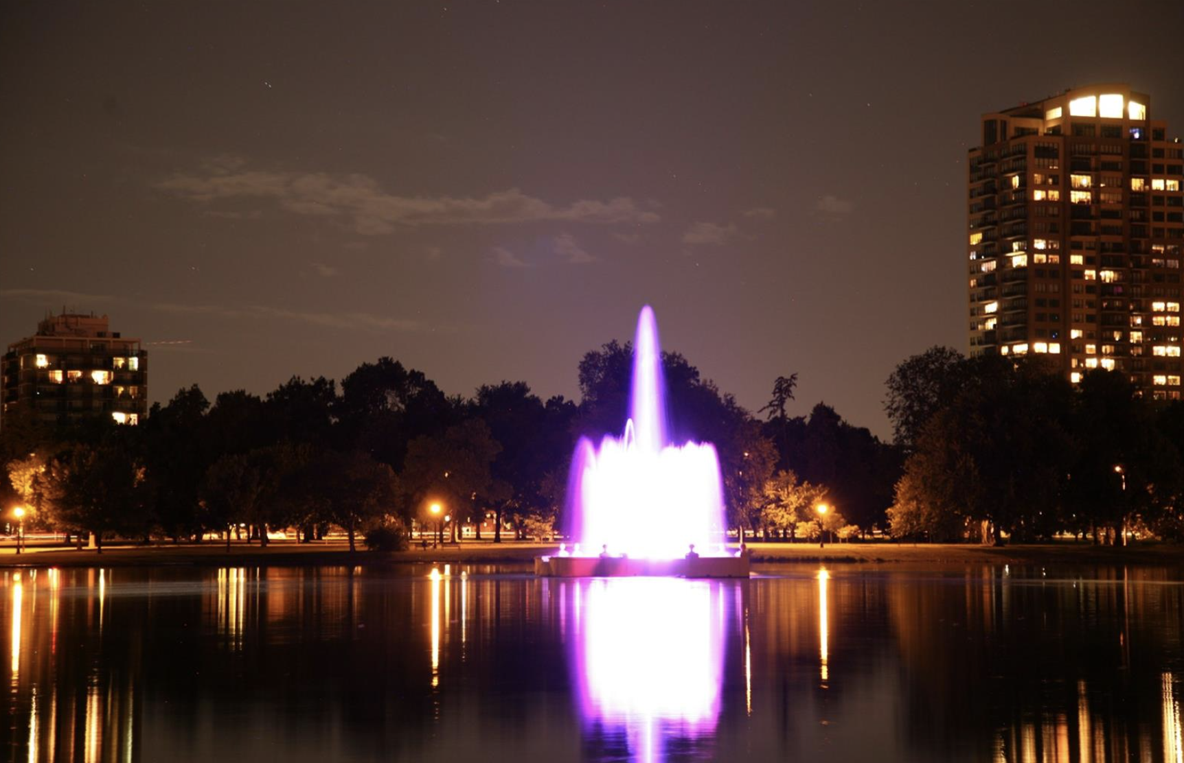 Nighttime scene of a brightly lit fountain in a park reflecting in a calm pond, with trees and tall residential buildings in the background at City Park, Denver, CO.