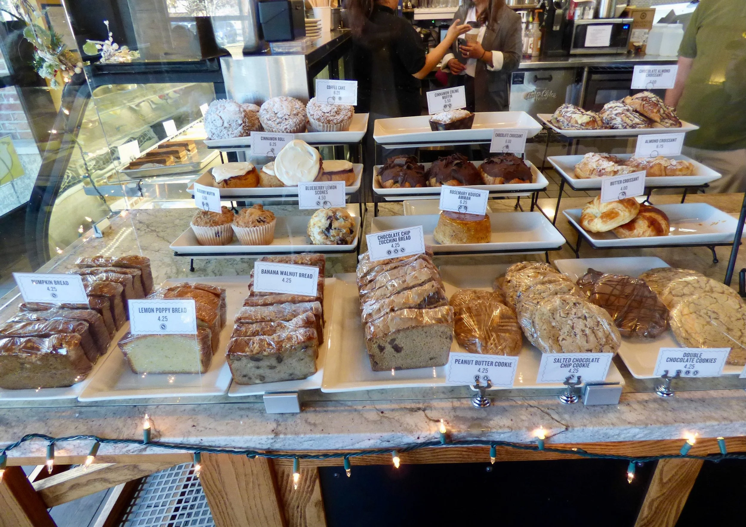 Display case with various baked goods including pumpkin bread, lemon poppy bread, banana walnut bread, cookies, muffins, croissants, and cakes in a bakery or coffee shop at Olive & Finch, Denver, CO.