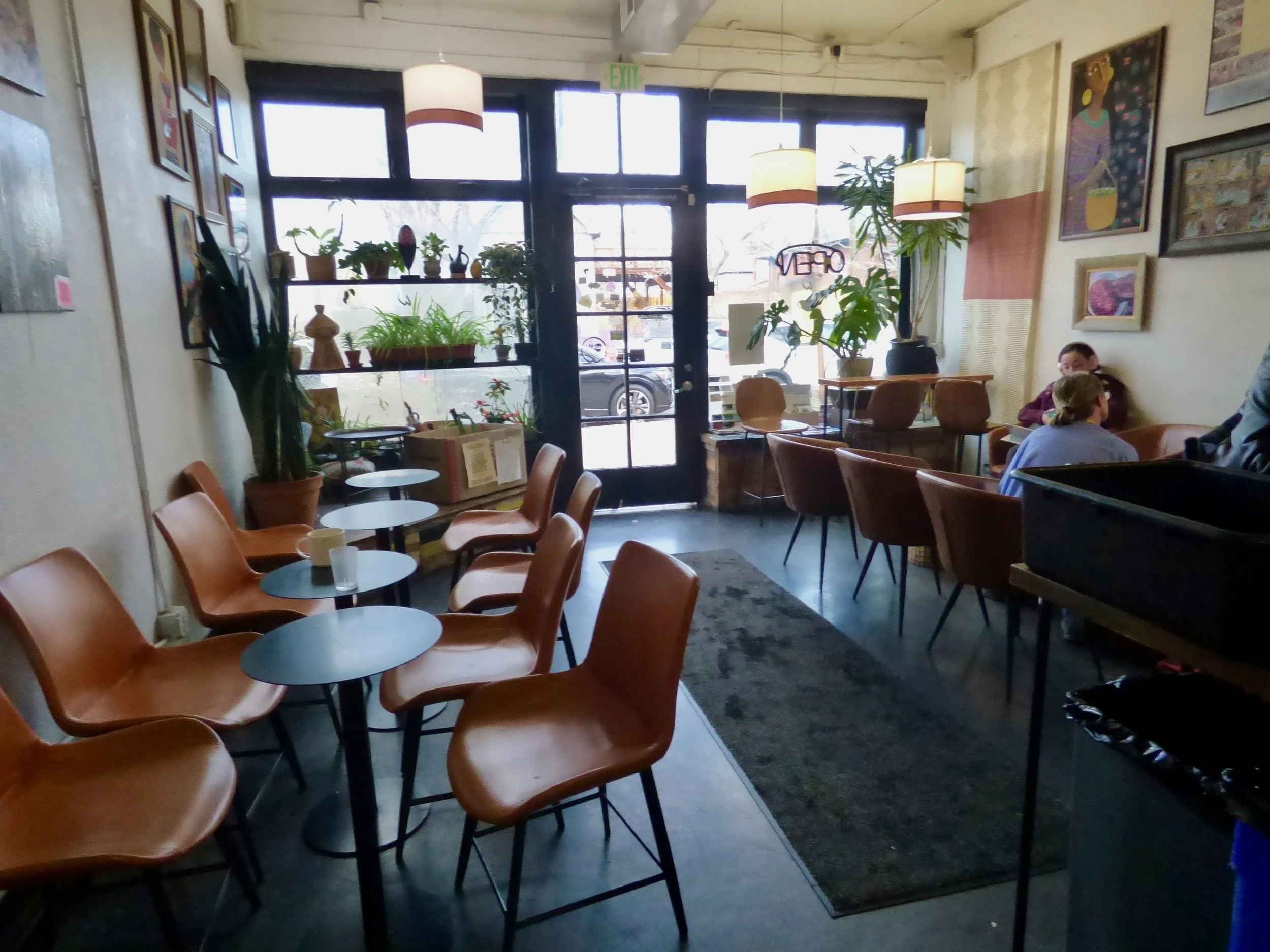 Interior of a cozy coffee shop with orange chairs, small circular tables, green plants, artwork on the walls, and a large window letting in natural light at Whittier Cafe, Denver, CO 80205.