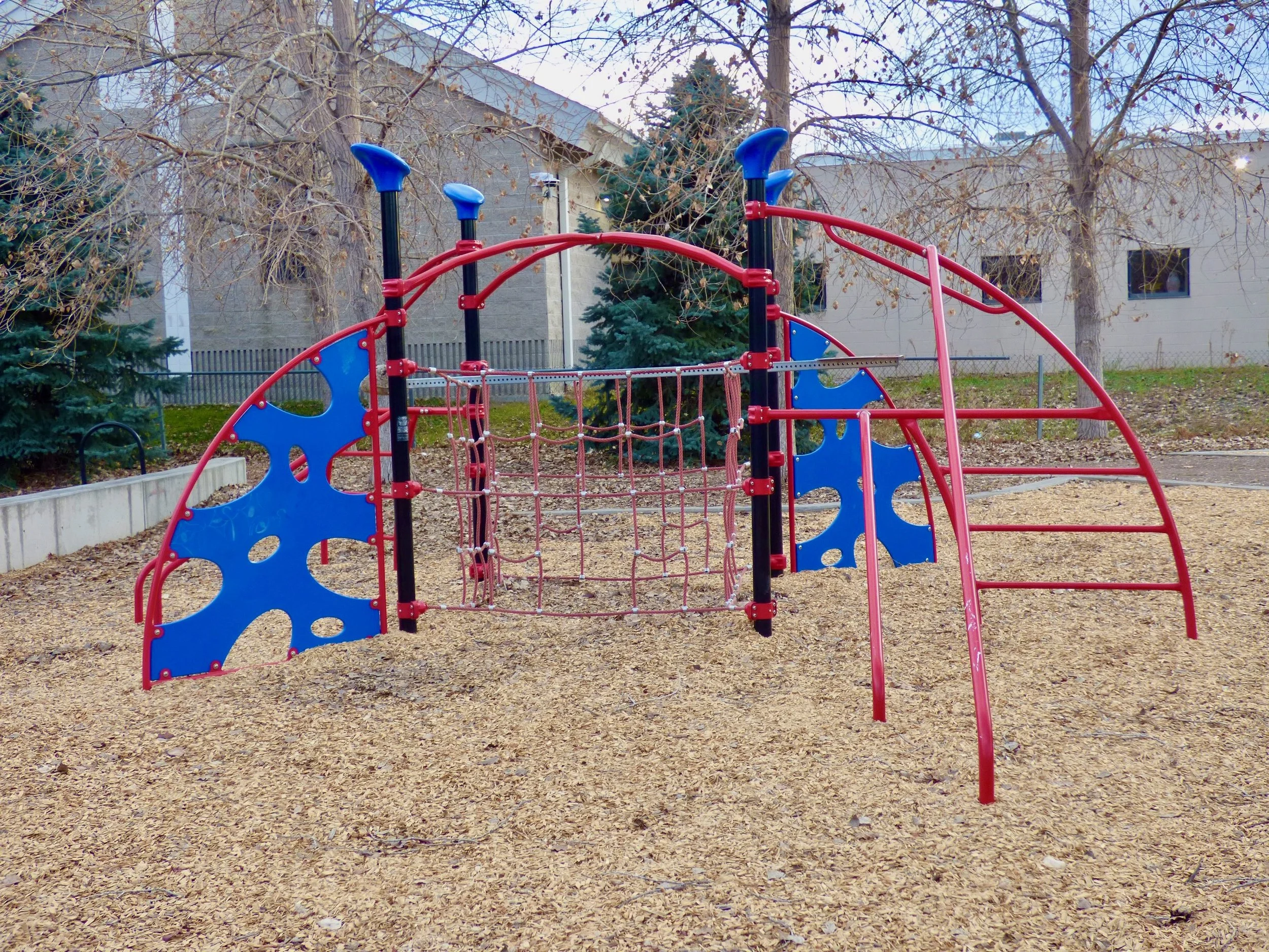 A colorful playground structure with red, blue, and black components in a park with trees and a building in the background at Madame C.J. Walker Park, Denver, CO 80205.