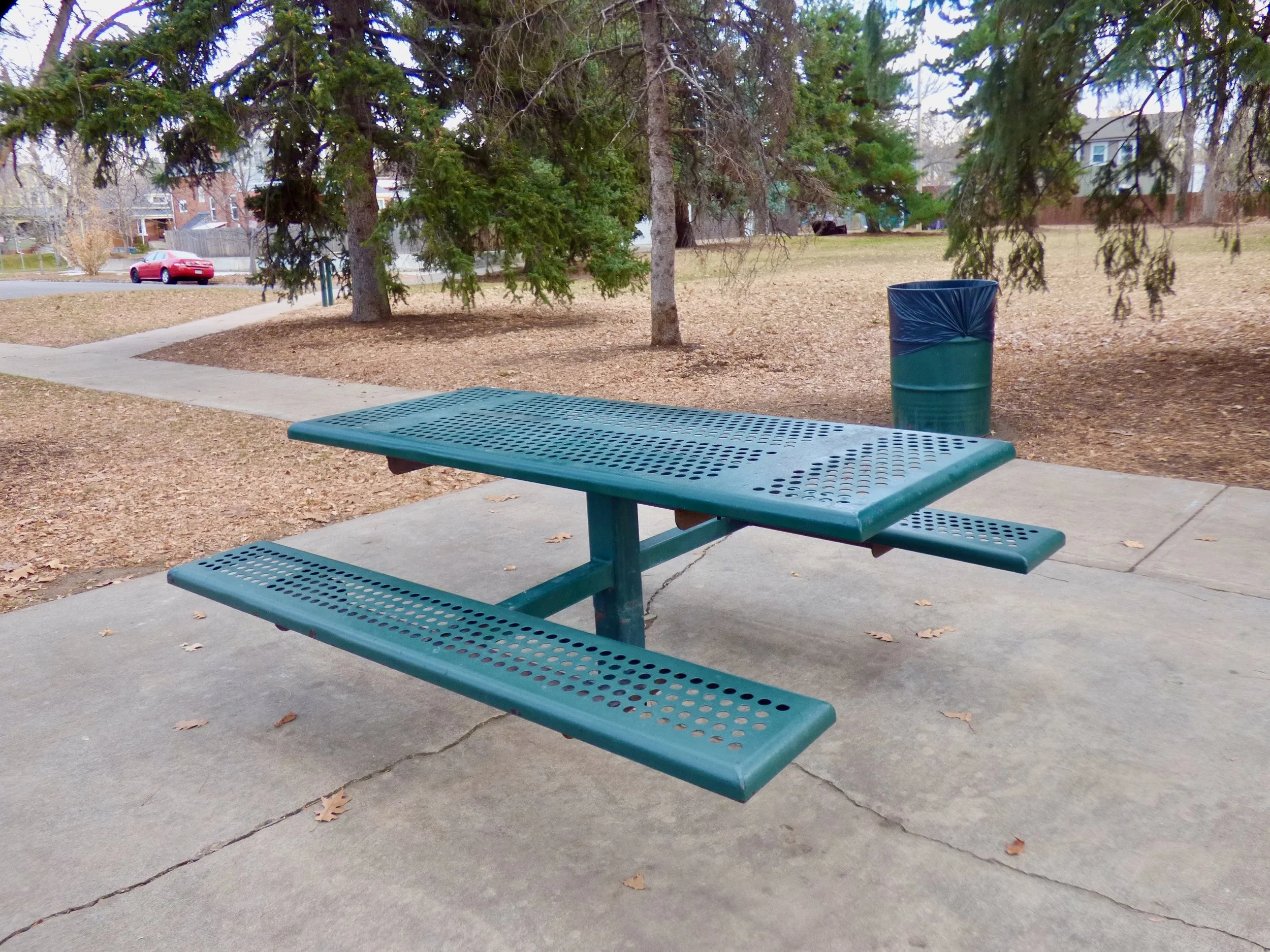 A green perforated metal picnic table with attached benches on a concrete sidewalk in a park, with trees and a metal trash can covered in a blue plastic bag in the background, during an overcast day at George Morrison Sr. Park, Denver, CO 80205.