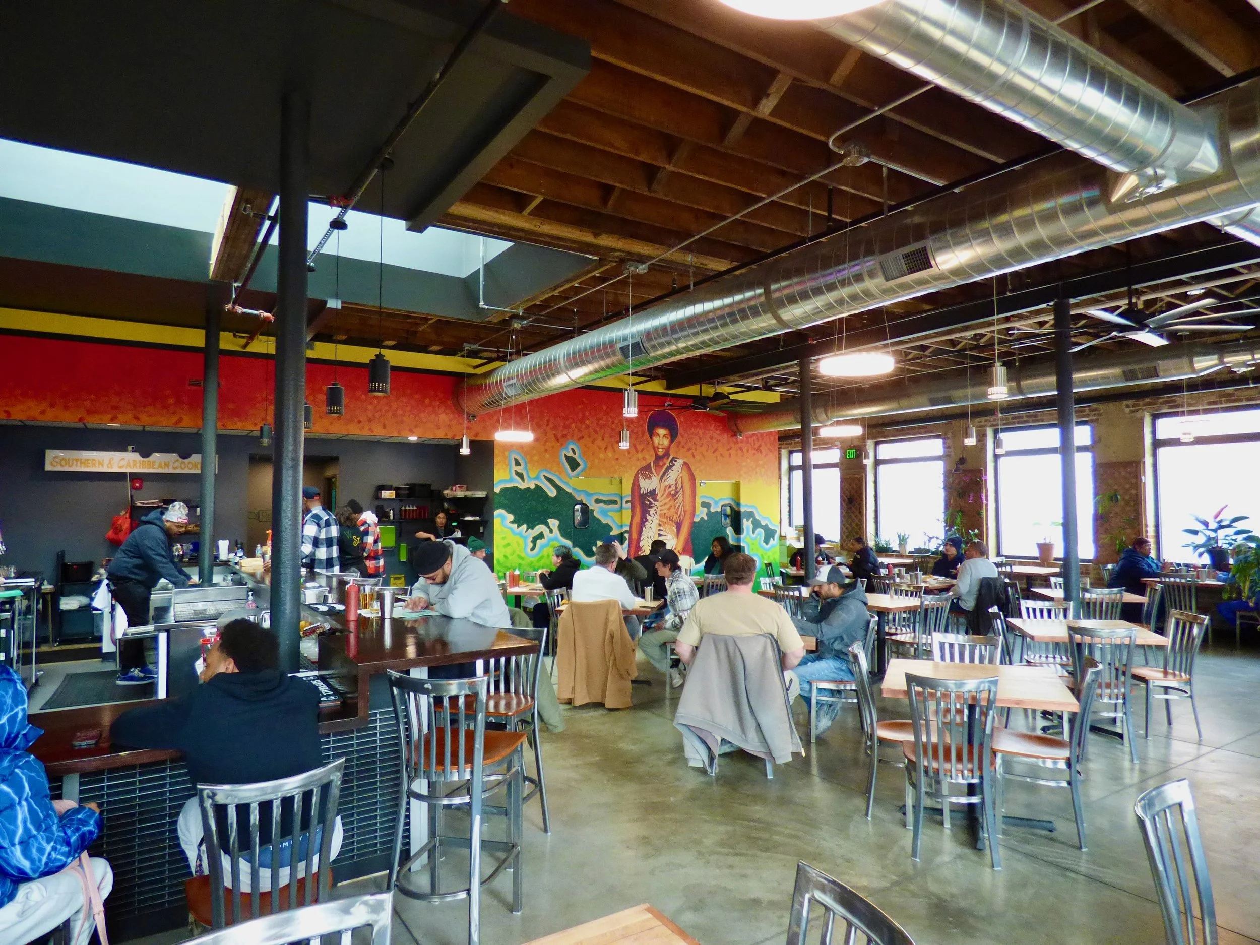 Interior of a busy restaurant with people sitting at tables and at the counter, colorful mural on the wall, large windows, and exposed ceiling pipes.