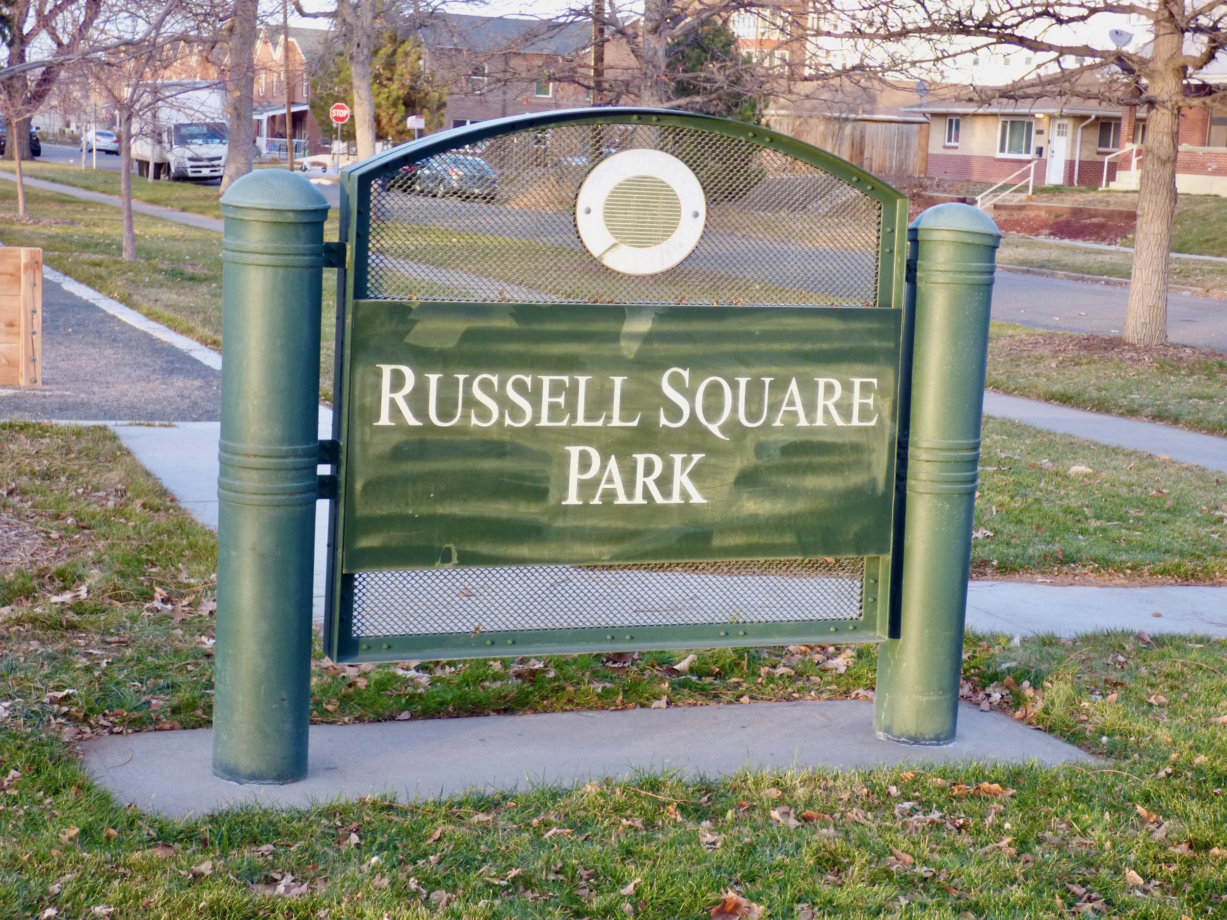Green park sign with white lettering that reads "Russell Square Park" situated on a sidewalk lawn, with trees, houses, and cars in the background in Russell Square Park, Denver, CO 80205.
