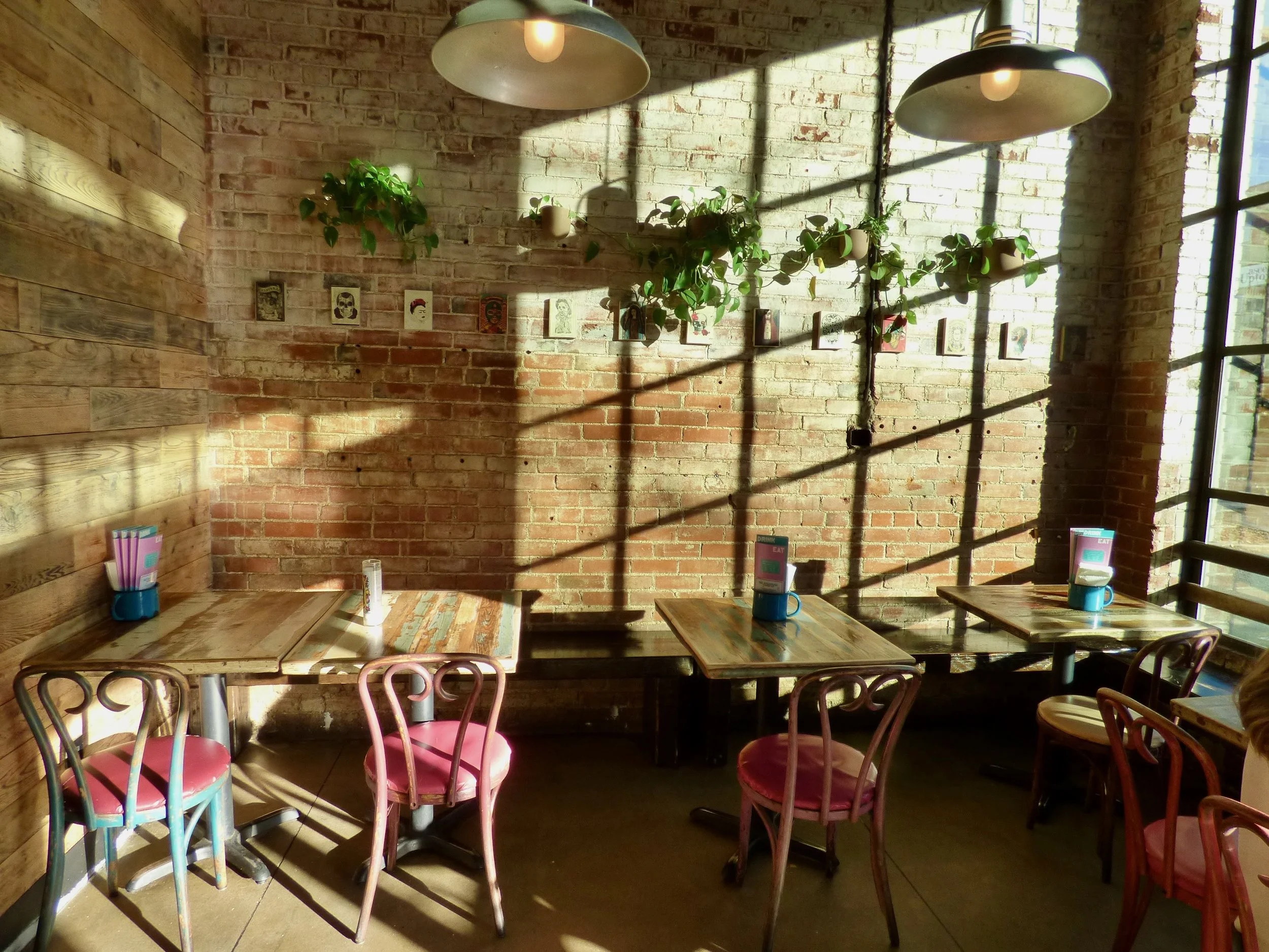 Sunlit corner of a restaurant interior with exposed brick walls, wooden tables, pink chairs, green potted plants, wall art, and large windows casting shadows at Dos Santos, Denver, CO.