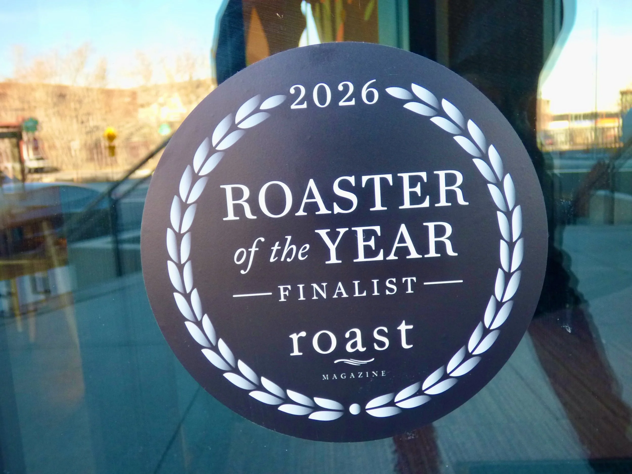 A circular sticker on a glass window reads "2026 Roaster of the Year Finalist roast" with a laurel wreath design around the text at Corvus Coffee Roasters, Denver, CO 80205.