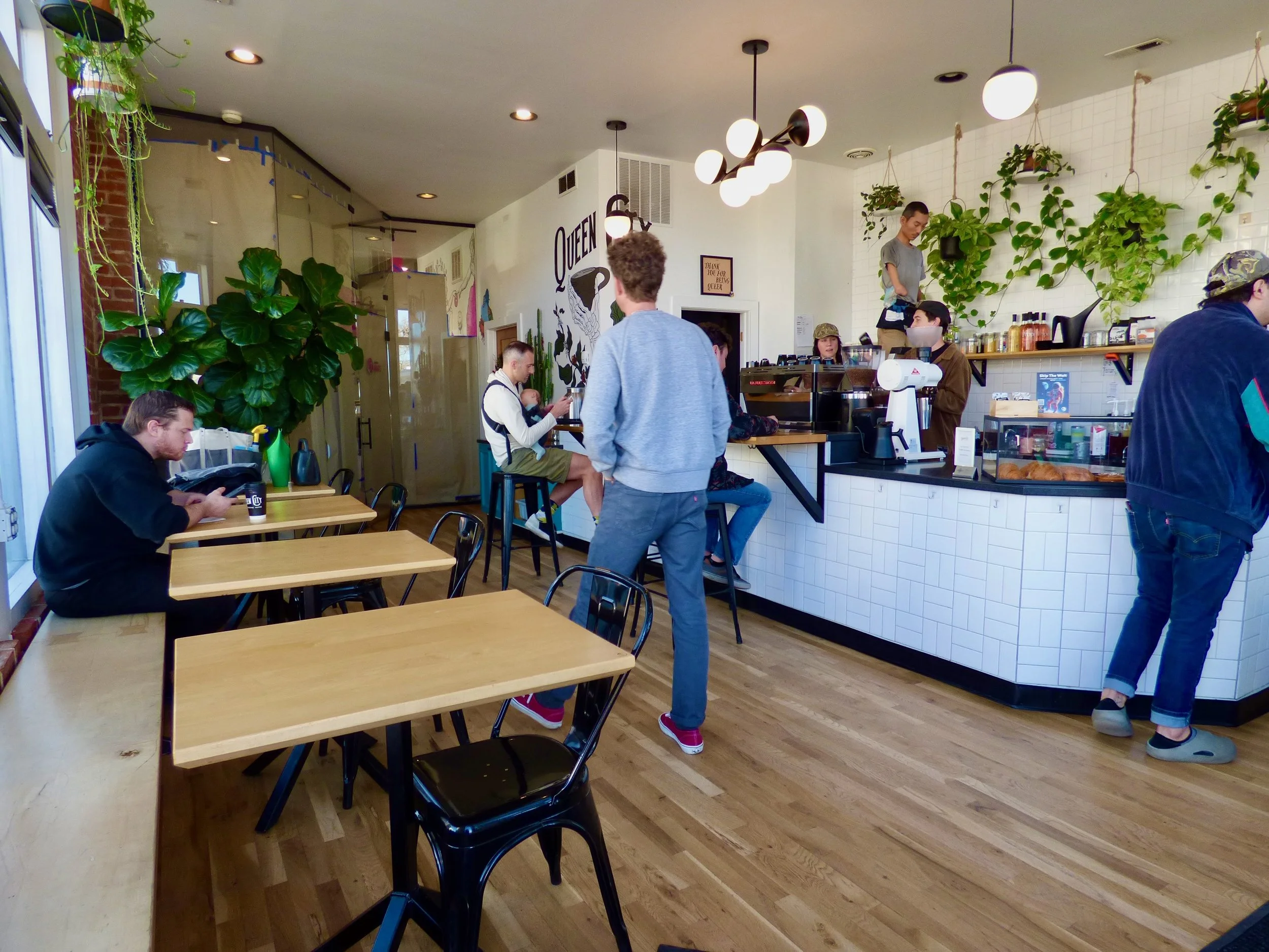 Inside a cozy coffee shop with wooden tables, green plants, and a counter where baristas prepare drinks. Customers are sitting and standing, some using phones at Queen City Collective Coffee House, Denver, CO 80205.