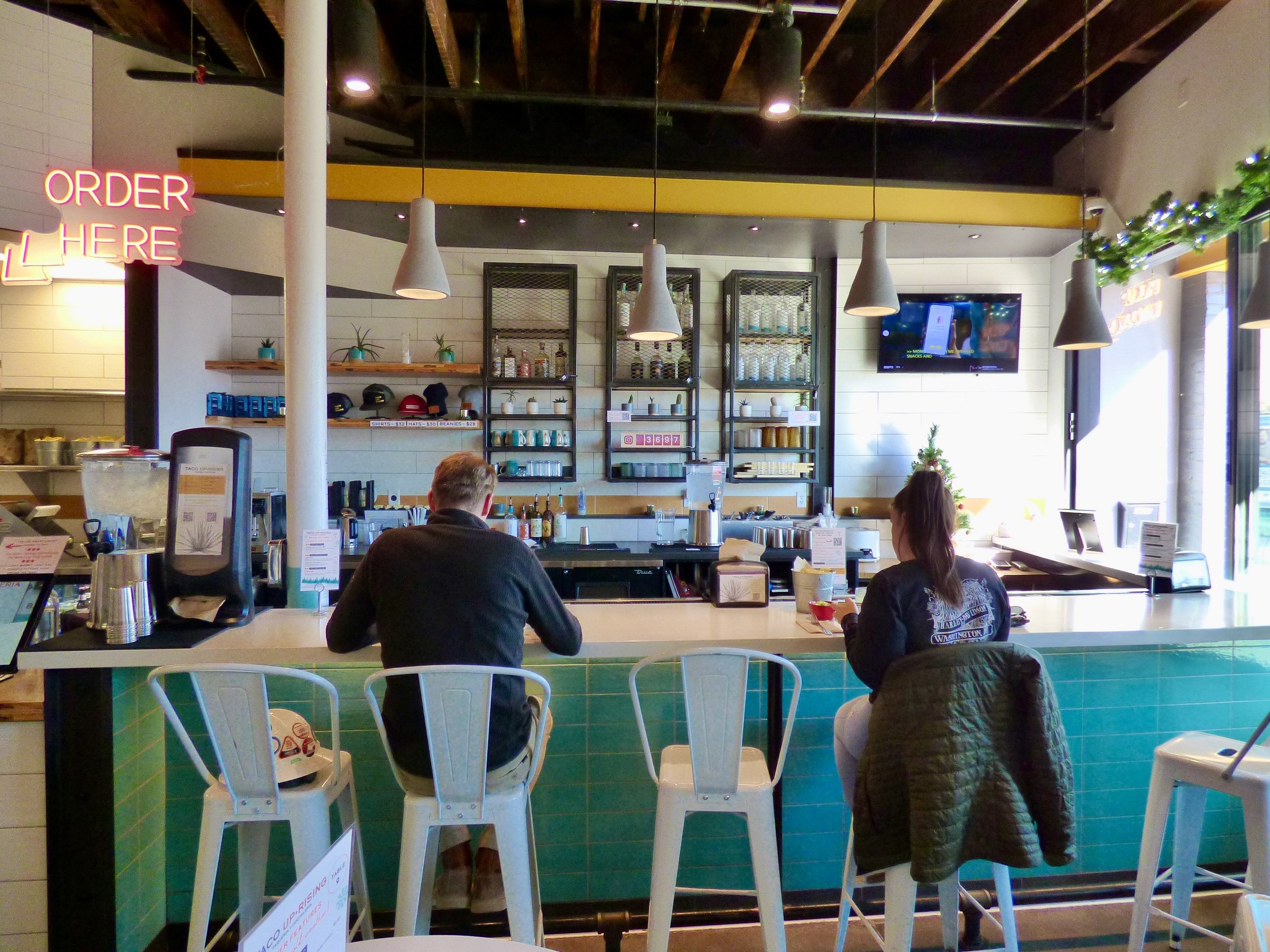 Inside a modern coffee shop with two customers sitting at the counter, one male and one female, with shelves of cups and bottles behind the bar, and a TV mounted on the wall.