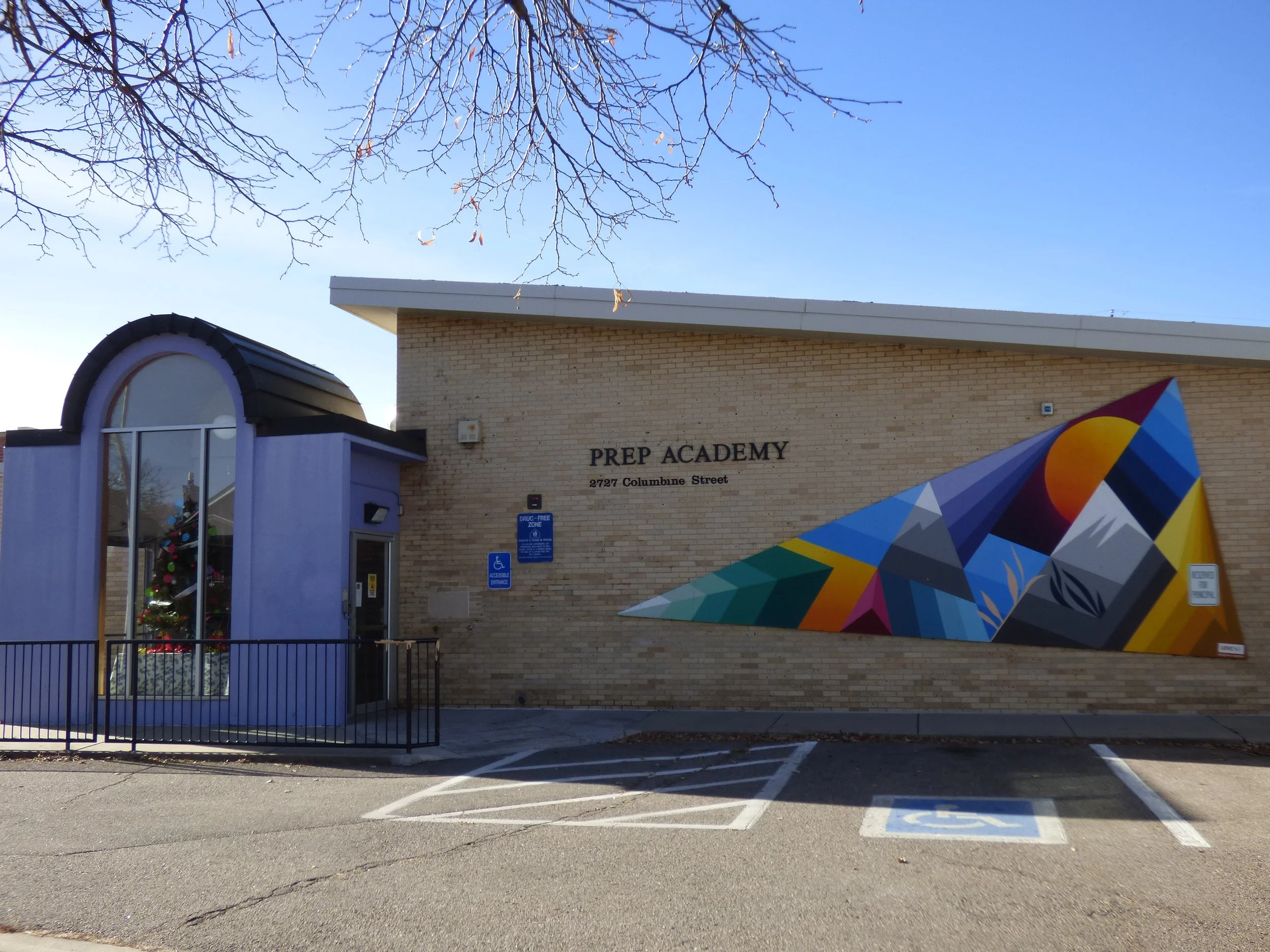 Brick building with sign reading 'Prep Academy, 2727 Columbine Street' and a colorful geometric mural on the right side. A small purple entrance with a curved glass window/display area, Christmas decorations visible inside. Handicap parking spaces with blue signs and painted symbols are in front, with leafless tree branches above, indicating winter.