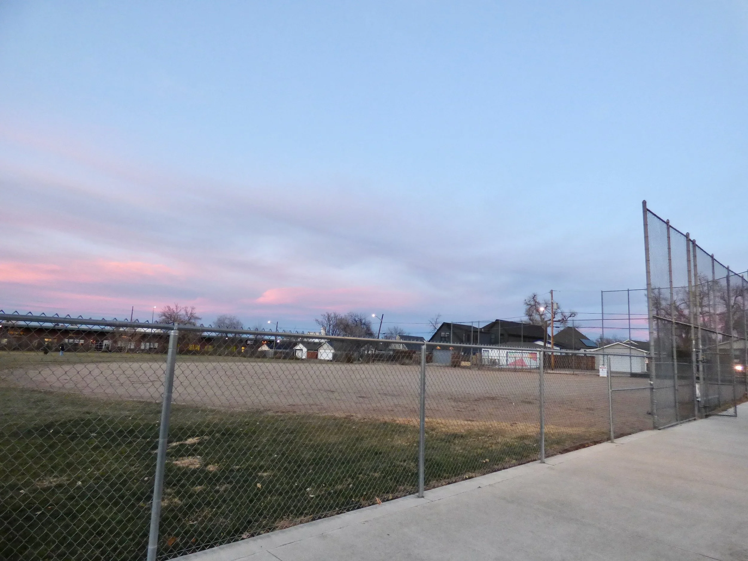 An empty baseball field with a chain-link fence around it, houses in the background, and a pink and blue sunset sky in Martin J. Schafer Park, Denver, CO 80205.