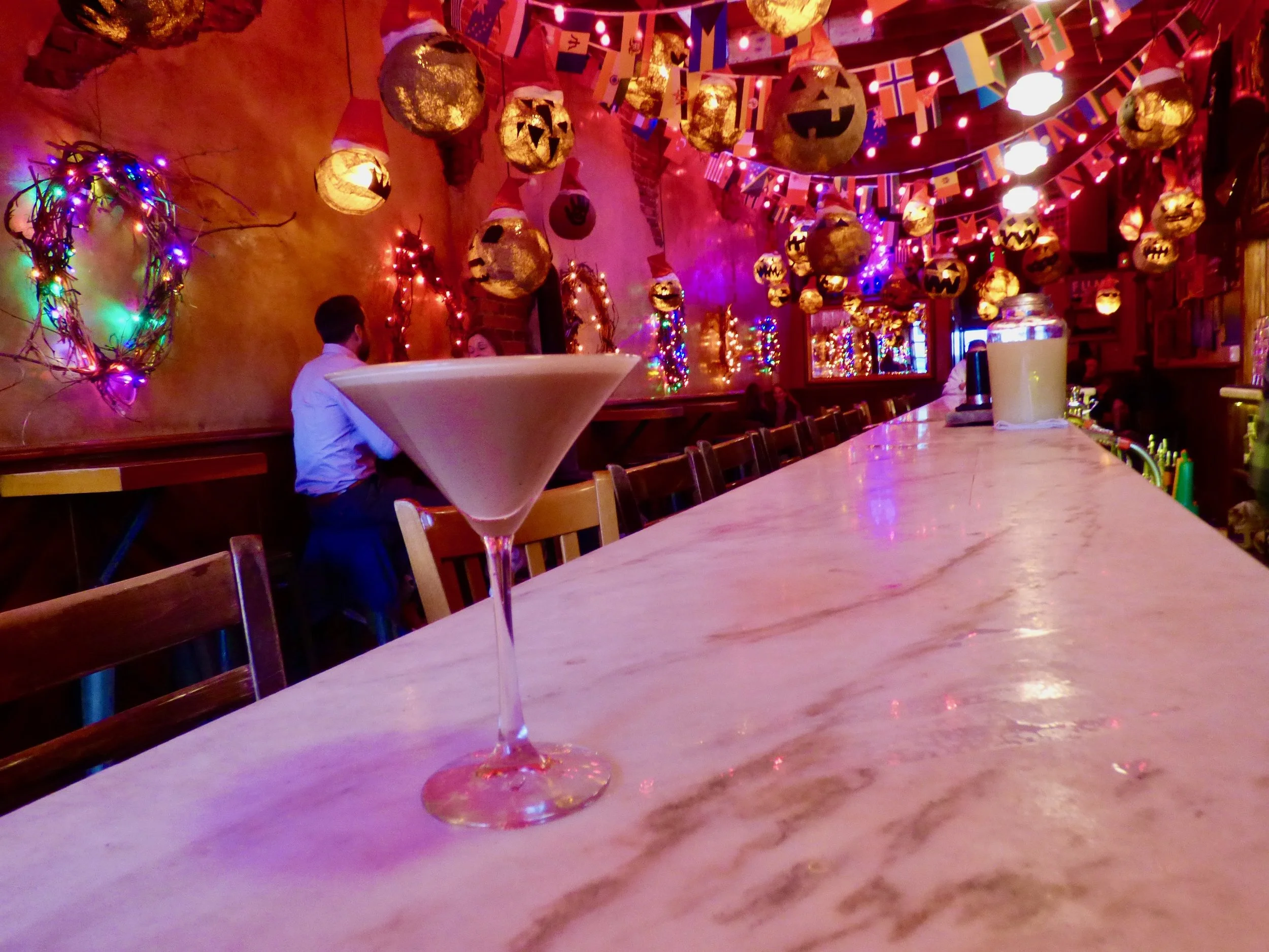 A martini glass on a bar counter in a dimly lit bar decorated with Halloween and Christmas ornaments, including carved pumpkin lanterns hanging from the ceiling, colorful string lights, and flags at The Thin Man, Denver, CO.