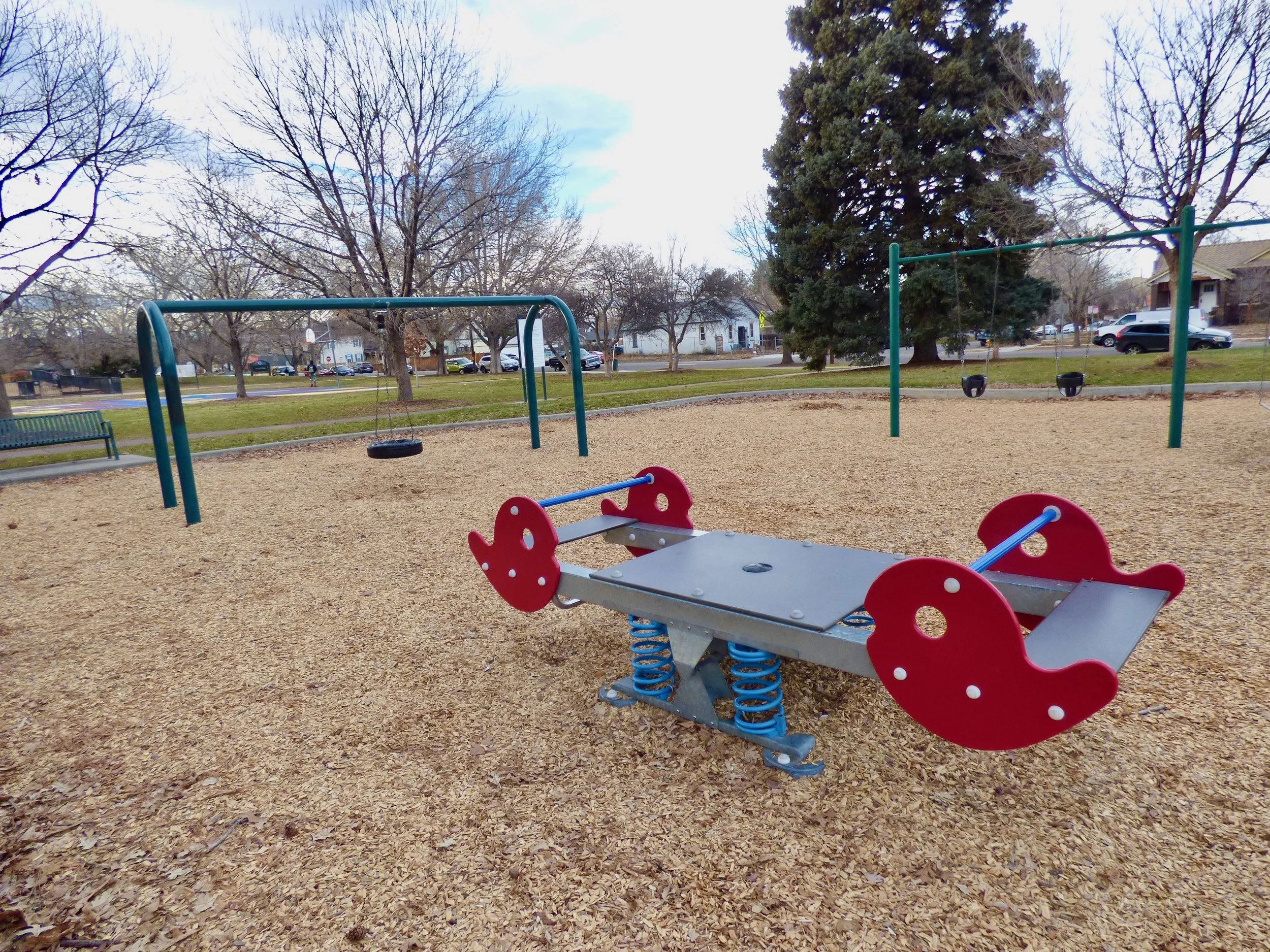 Playground with swings, see-saw, and spring rider in a park, no children present, leafless trees and houses in background at Fuller Park, Denver, CO 80205.