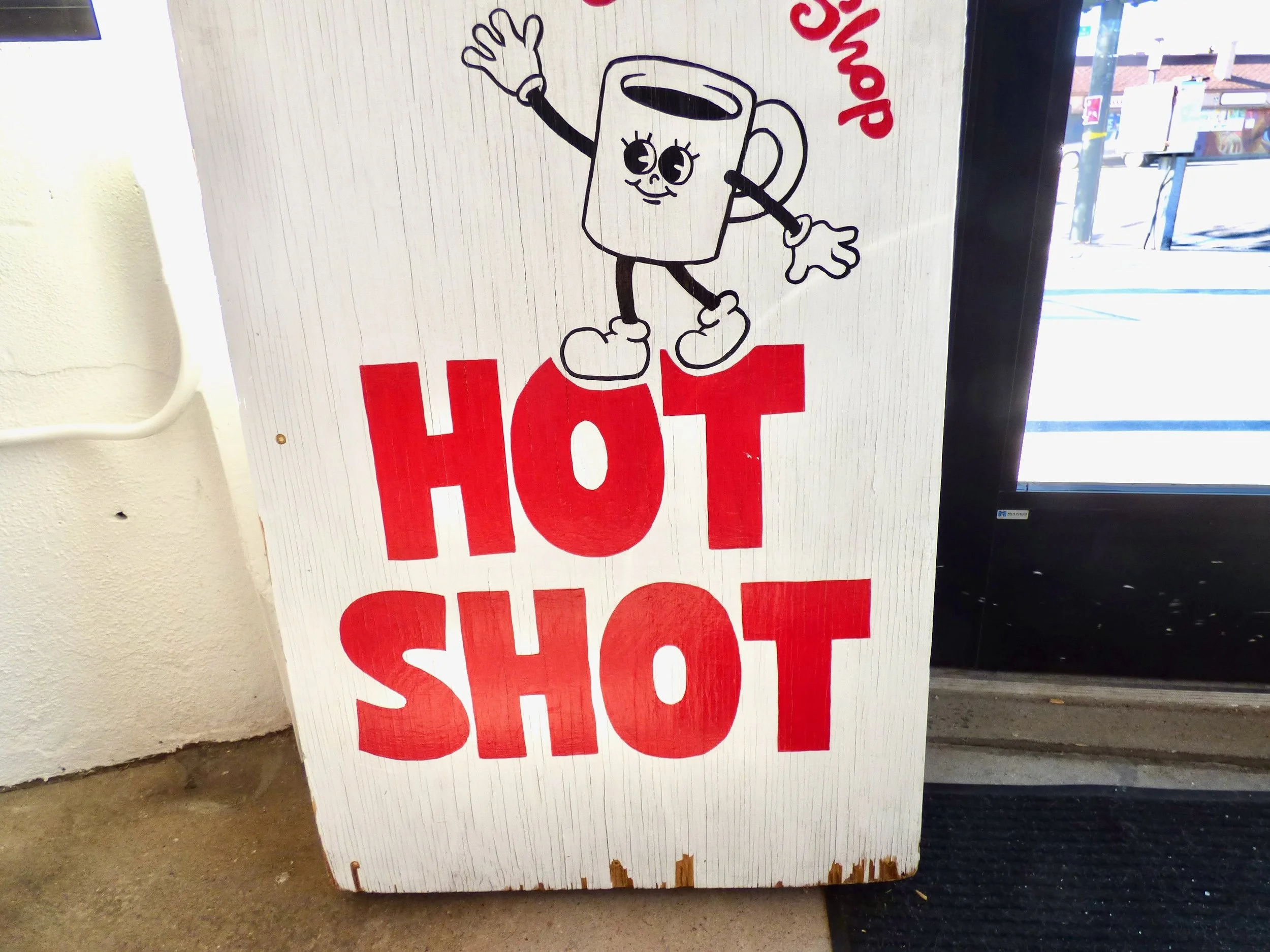 A white wooden sign with a cartoon coffee mug character waving, above the words "Hot Shot" in bold red letters at Hot Shot Coffee Shop, Denver, CO 80205.