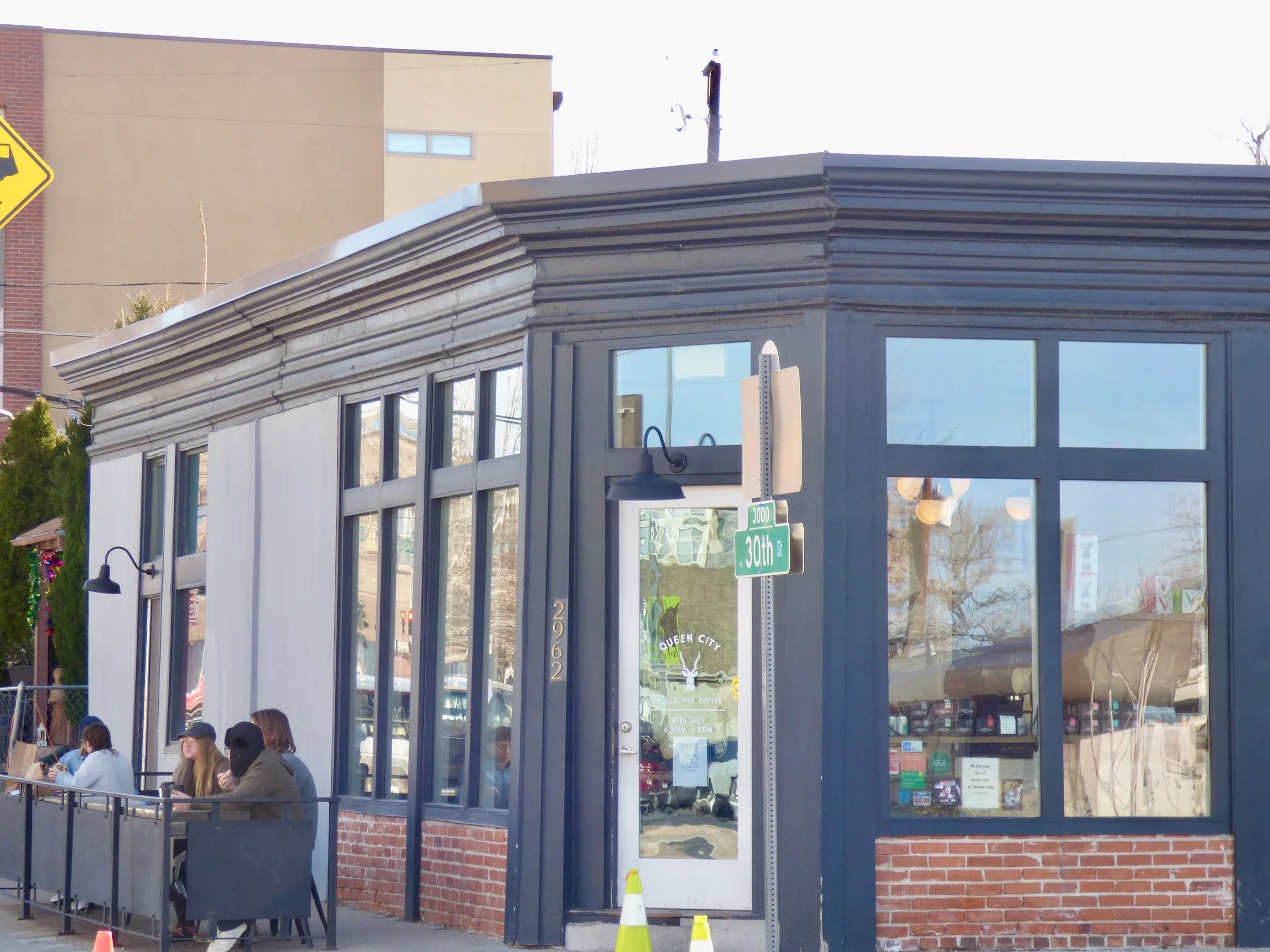 Exterior view of Queen City Collective Coffee shop at 3000 30th Street, with a few customers sitting outside at tables, large windows, and signage at Queen City Collective Coffee House, Denver, CO 80205.