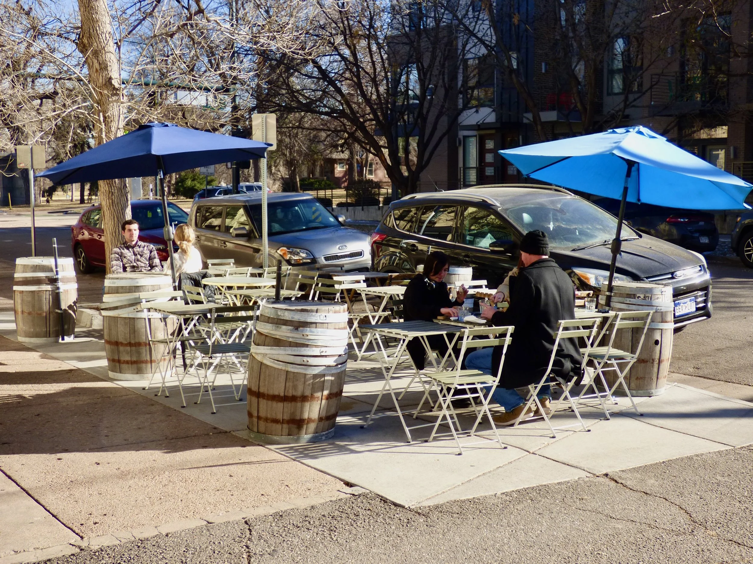 Outdoor sidewalk cafe with tables, chairs, and blue umbrellas in a city neighborhood, with parked cars and trees in the background at Cafe Miriam, Denver, CO 80205.