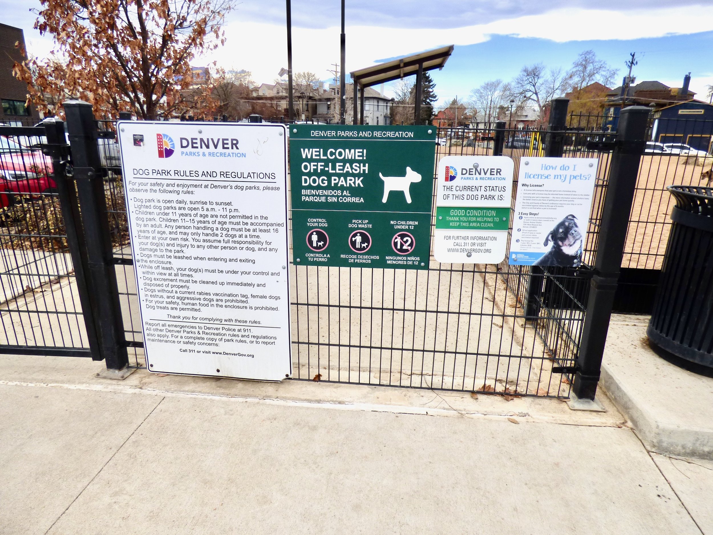 Entrance to a Denver parks and recreation off-leash dog park with multiple informational signs about dog rules, current park status, and pet licensing.