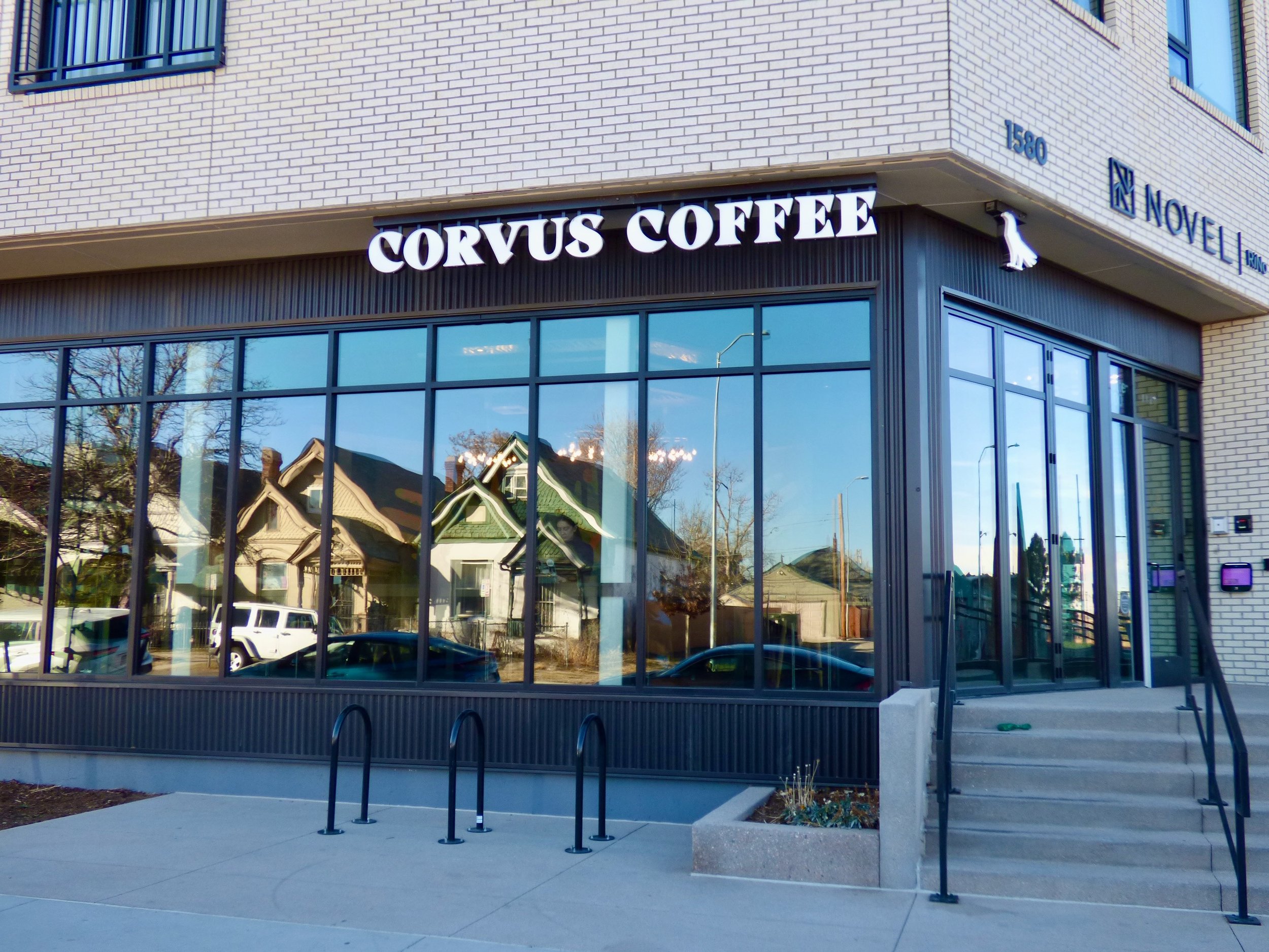 Exterior view of Corvus Coffee shop with large glass windows, black trim, and a sign above the entrance. Residential houses and parked cars reflect in the windows at Corvus Coffee Roasters, Denver, CO 80205.
