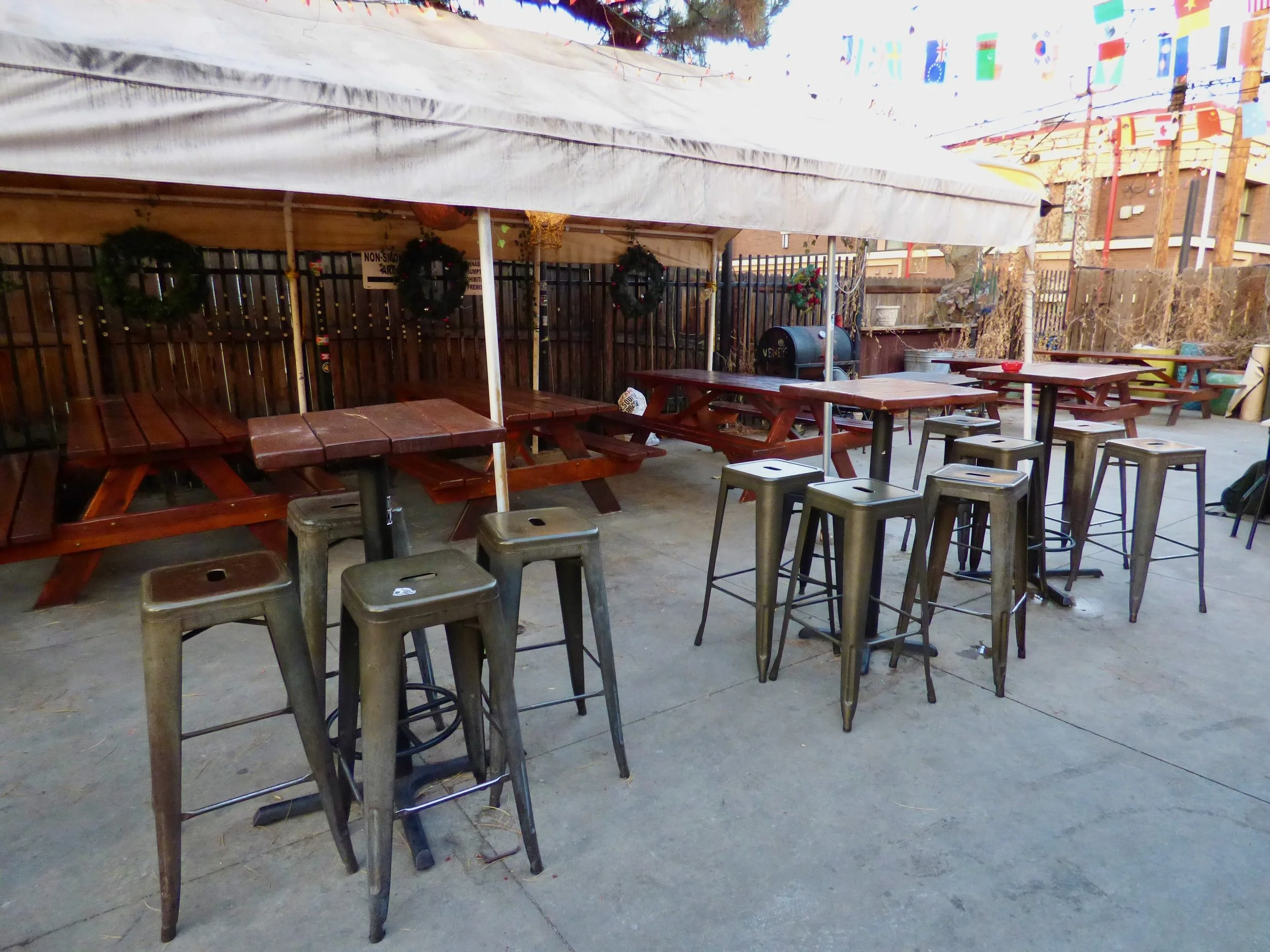 Empty outdoor patio with wooden picnic tables under a canopy, metal bar stools, holiday wreaths, and string lights at The Thin Man, Denver, CO.