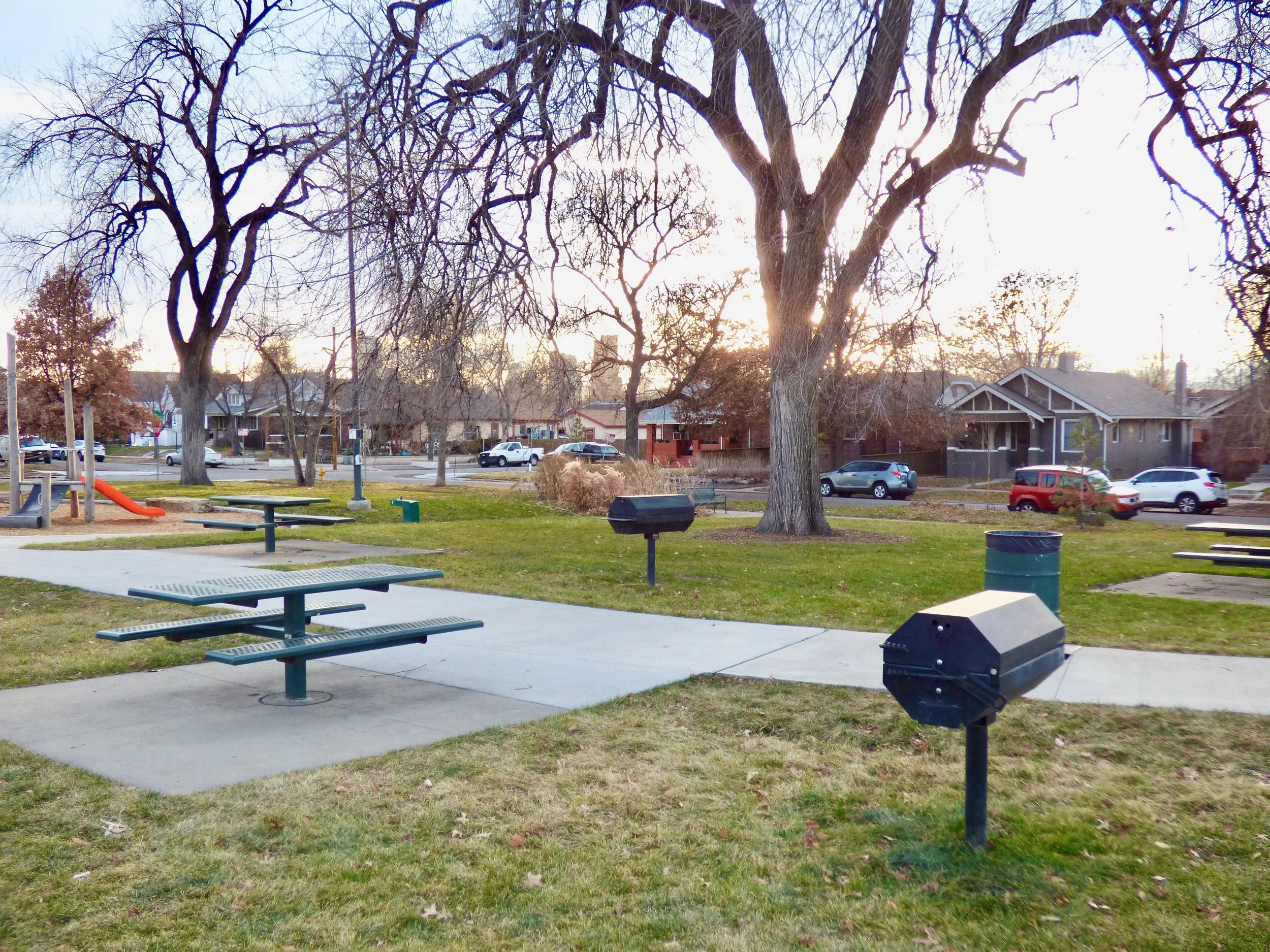 A park with picnic tables, barbecue grills, a trash can, and large leafless trees, with houses and parked cars in the background during sunset in Russell Square Park, Denver, CO 80205.