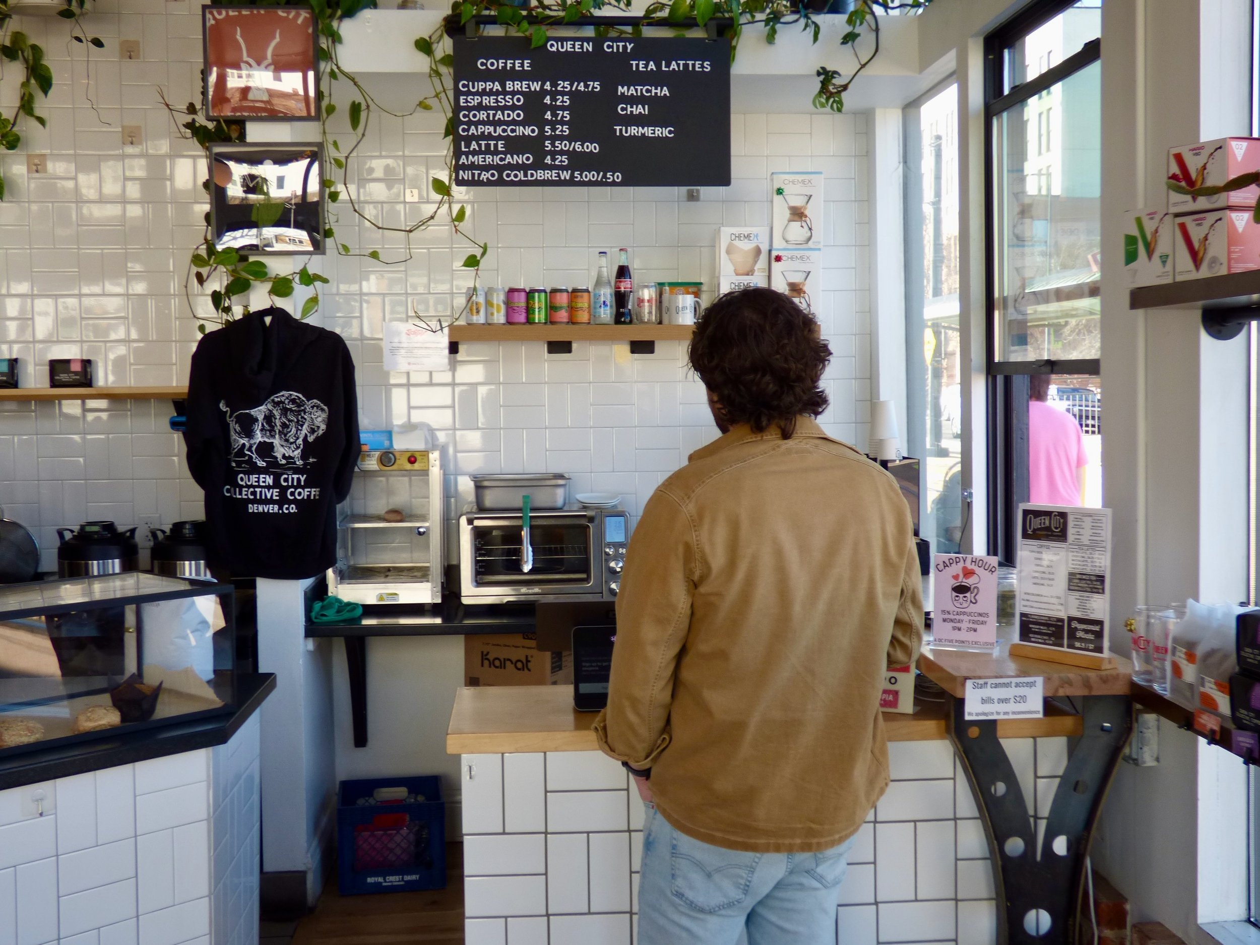 A person wearing a tan jacket stands at the counter of a coffee shop, facing away. The shop has white tiles on the wall, a black menu board listing coffee and tea options, and various supplies at Queen City Collective Coffee House, Denver, CO 80205.