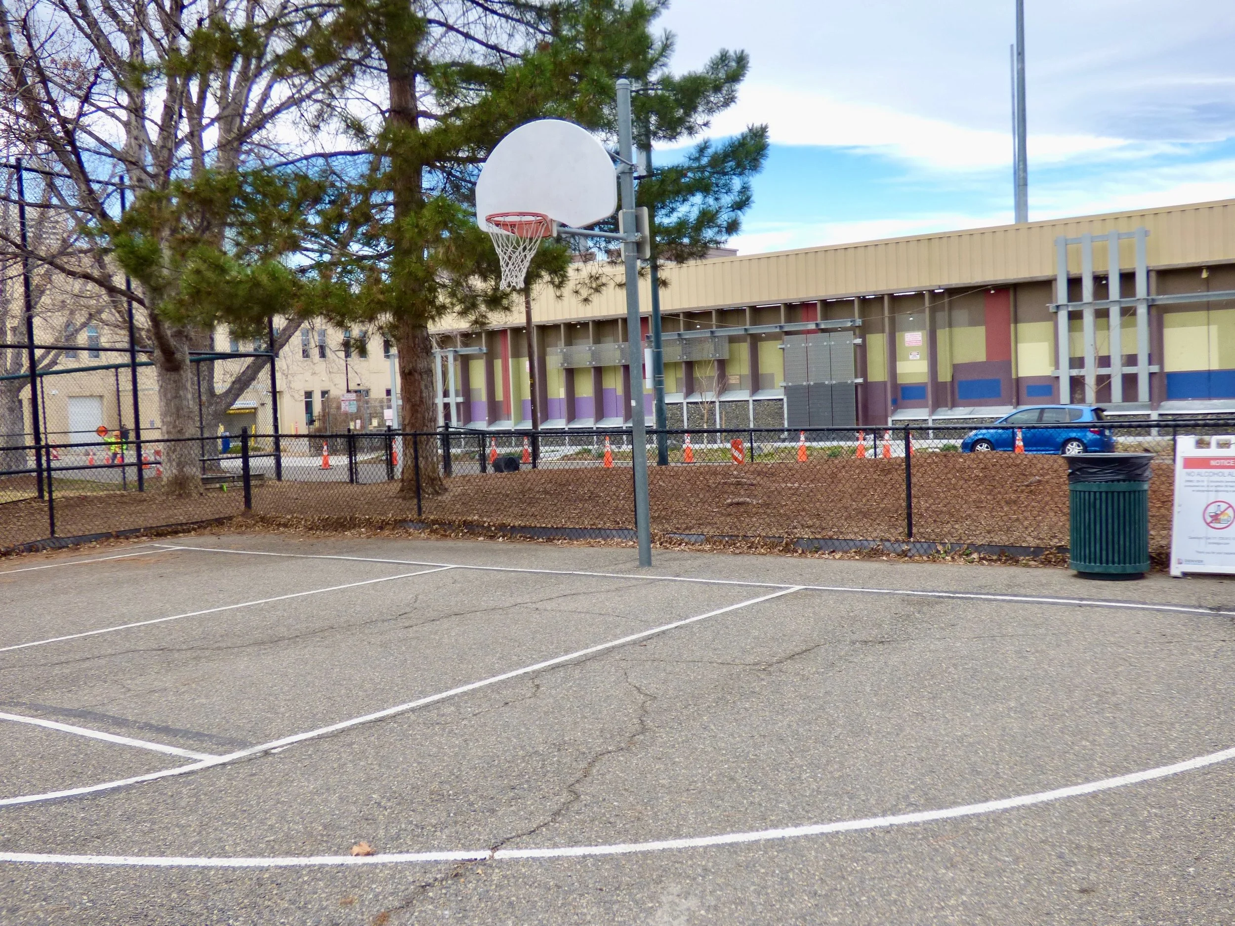Empty outdoor basketball court with a basketball hoop, surrounded by a black chain-link fence, with trees and a colorful building in the background in Sonny Lawson Park, Denver, CO 80205.