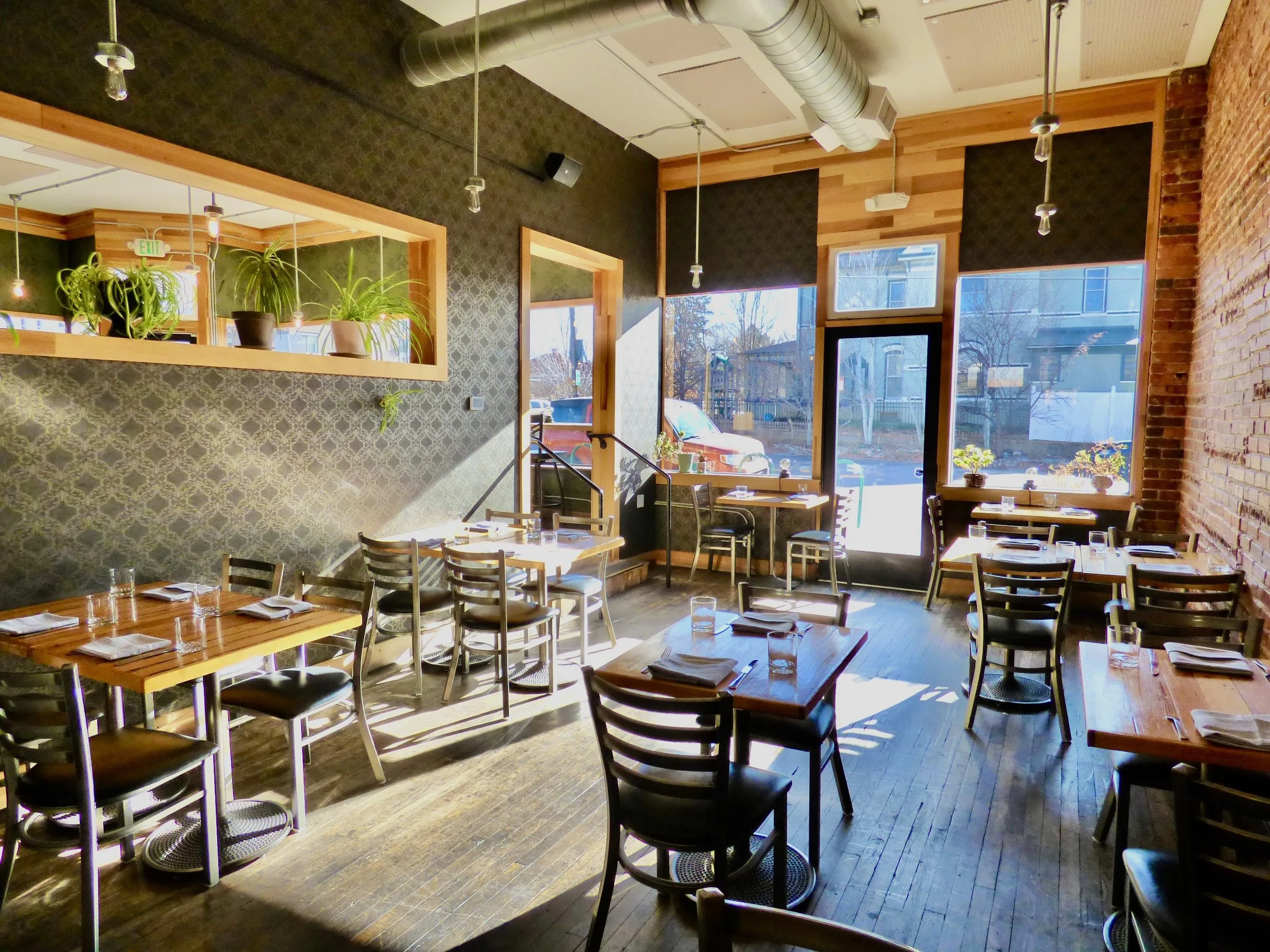 Inside a cozy restaurant with wooden tables and black chairs, large windows letting in sunlight, green plants on a shelf, exposed brick wall, and dark patterned wallpaper at The Plimoth, Denver, CO 80205.