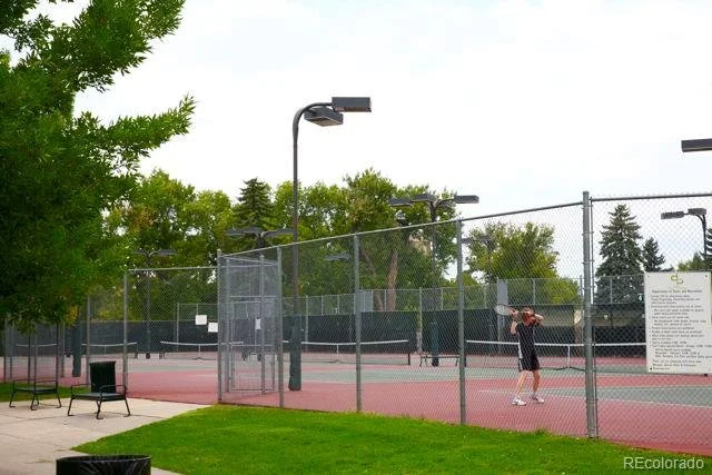 A person playing tennis on an outdoor court during the daytime, surrounded by a chain-link fence with trees in the background at City Park, Denver, CO.