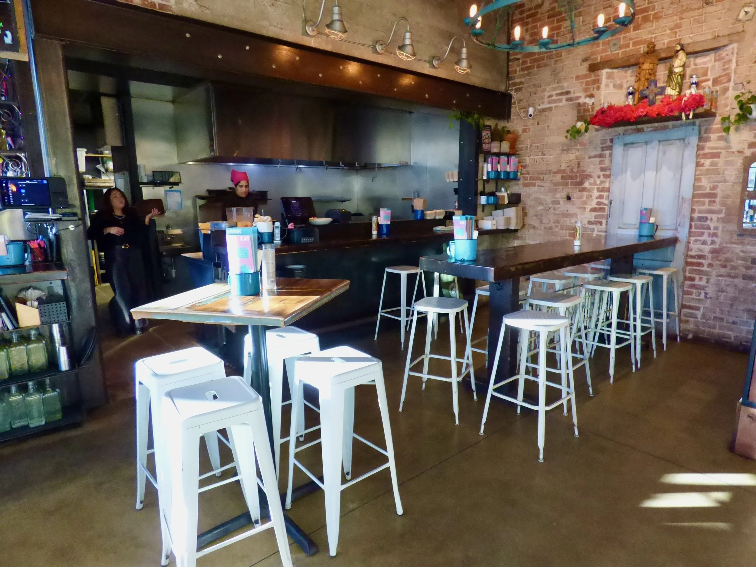 Interior of a coffee shop with high-top tables and bar seating, brick walls, and a kitchen area in the background with two staff members, one wearing a pink headscarf at Dos Santos, Denver, CO.