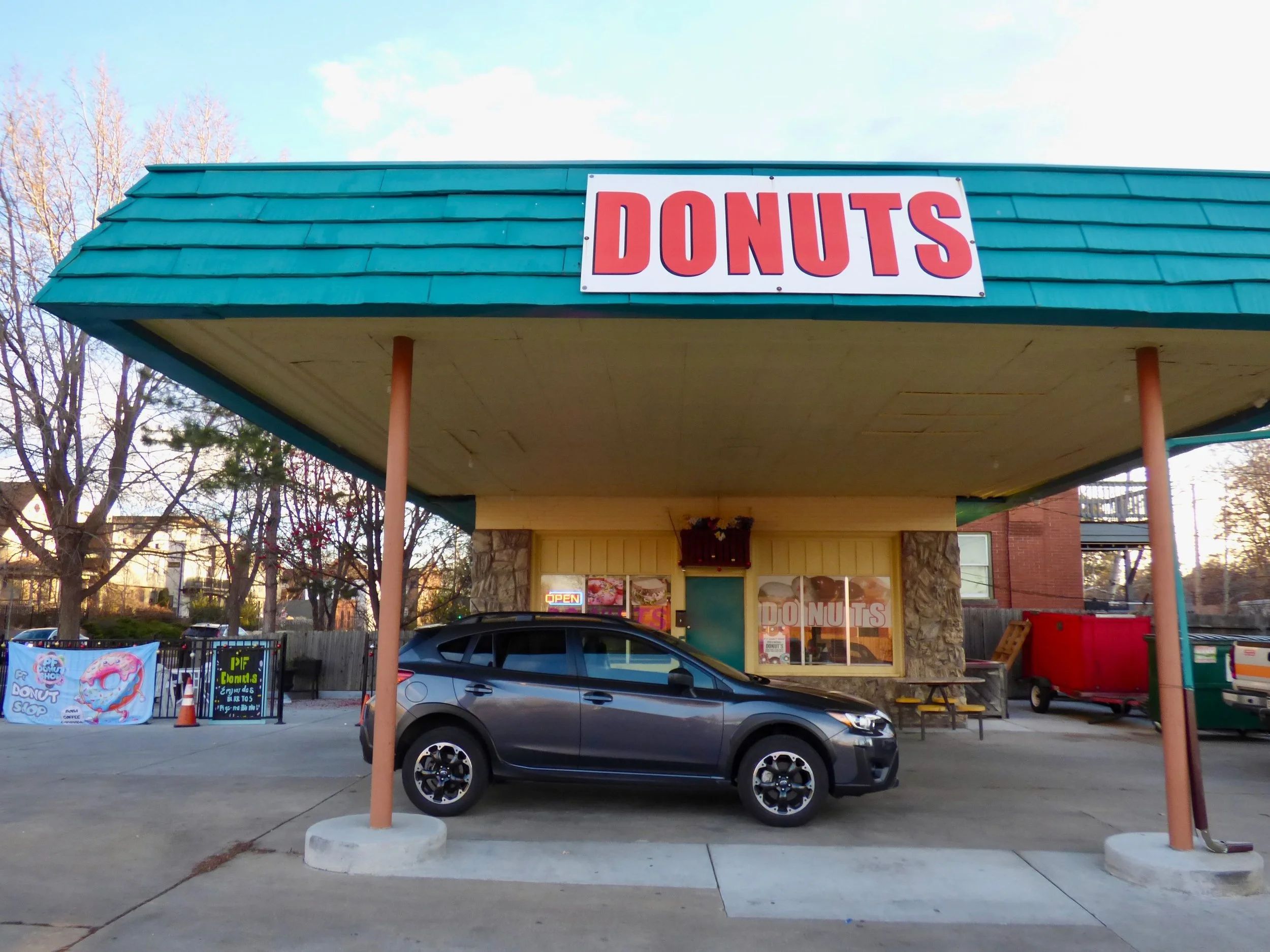 A fast food stand with a large red and white "DONUTS" sign on a teal roof, a black car parked in front, and trees in the background at PF Donut Shop, Denver, CO 80205.
