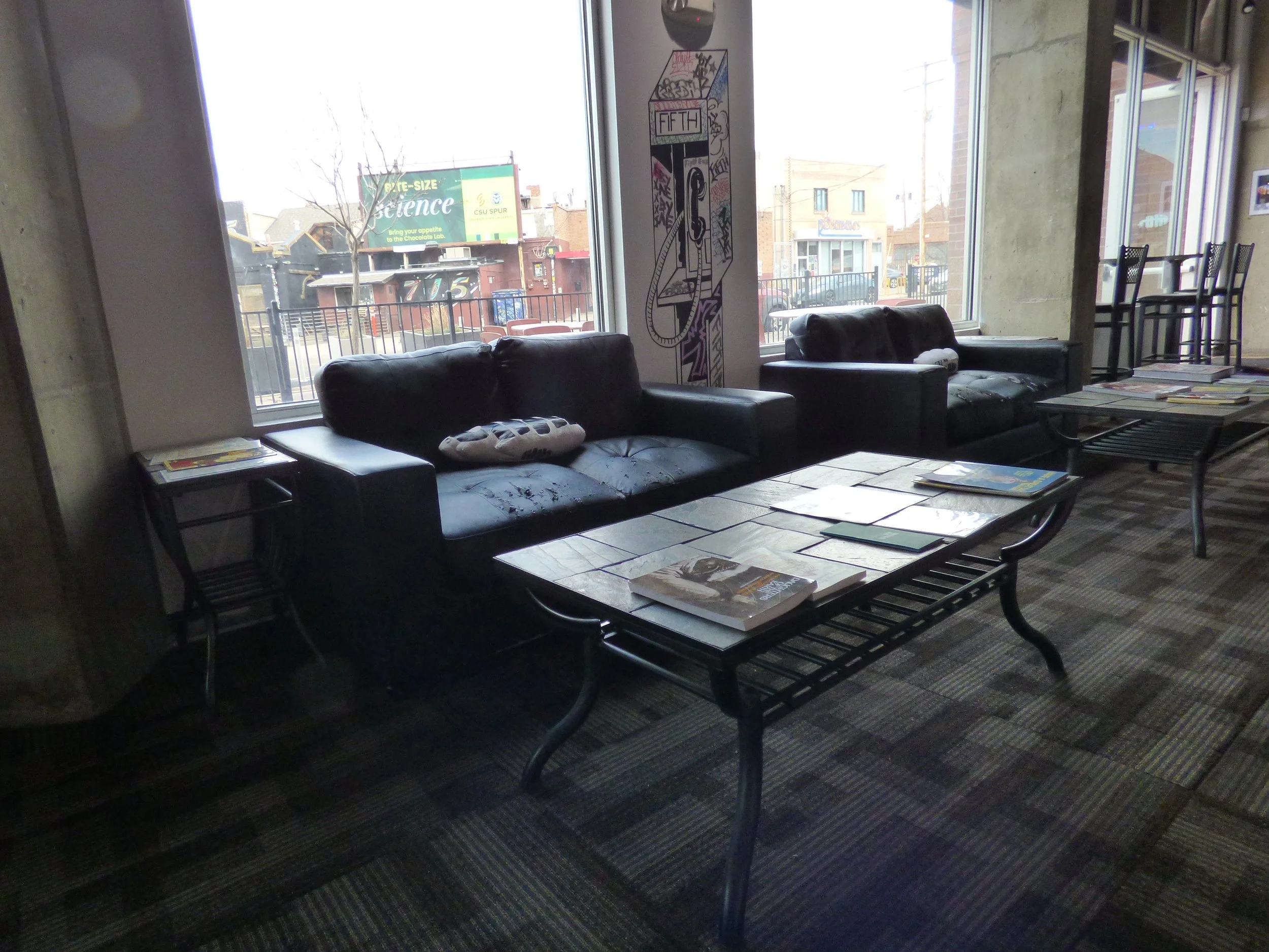 Indoor lounge area with black leather sofas, metal coffee tables with magazines, large windows, and a wall with artwork. Outside view shows buildings, a street, and a billboard Fifth Coffee House, Denver, CO 80205.