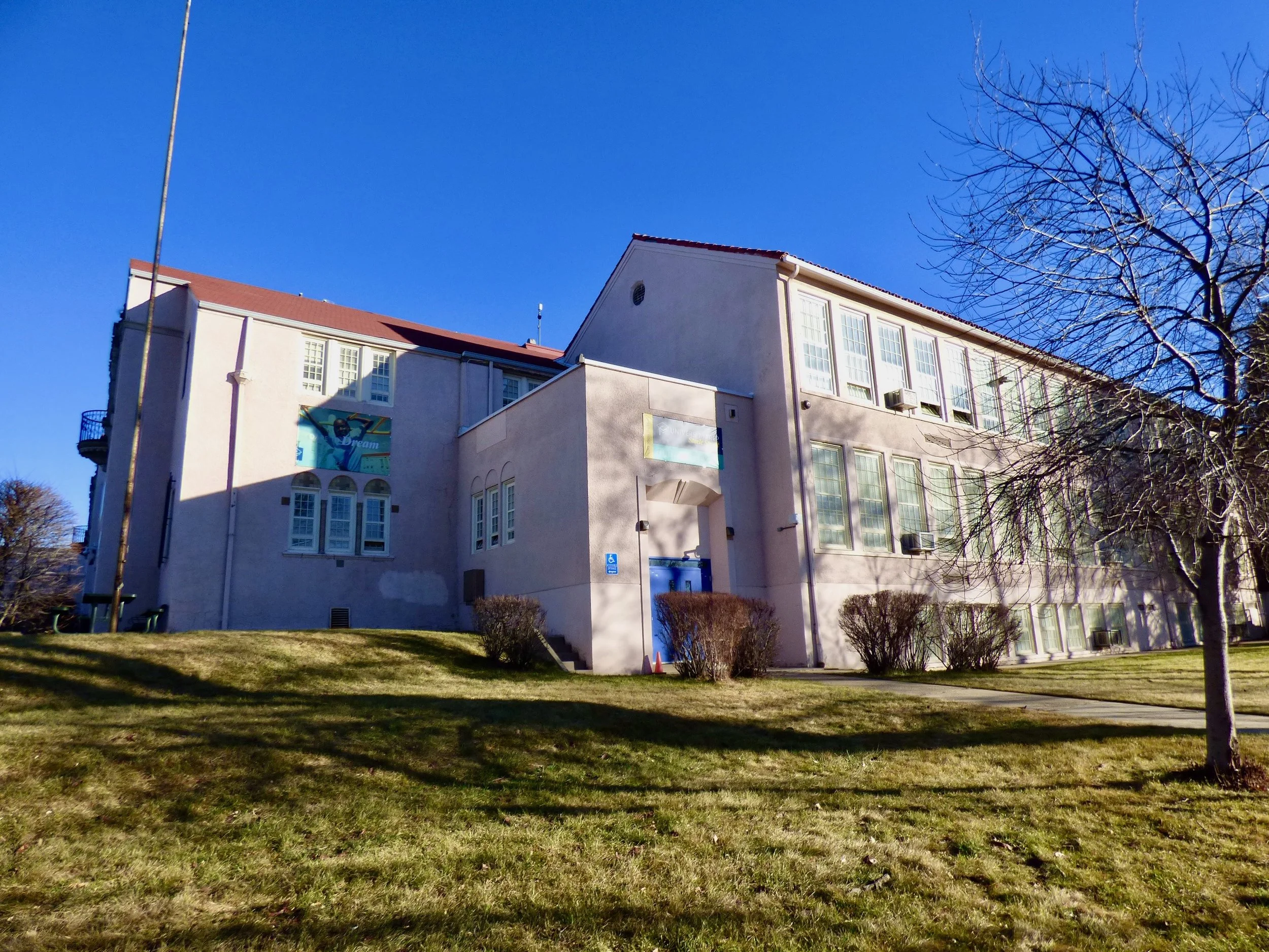 A multi-story beige building with large windows and a red roof, surrounded by a grassy yard with small bushes and a leafless tree, under a clear blue sky.