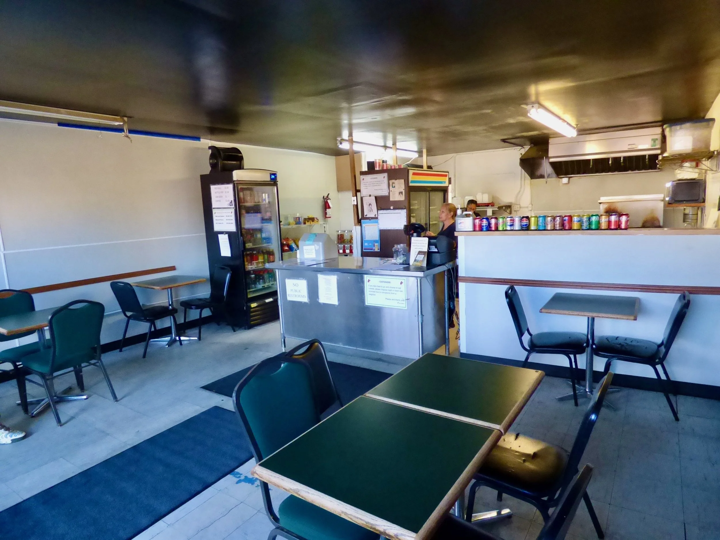 Interior of a small restaurant with empty tables and chairs, a counter with a cash register, a staff member standing behind it, a refrigerator, and canned drinks on a shelf.
