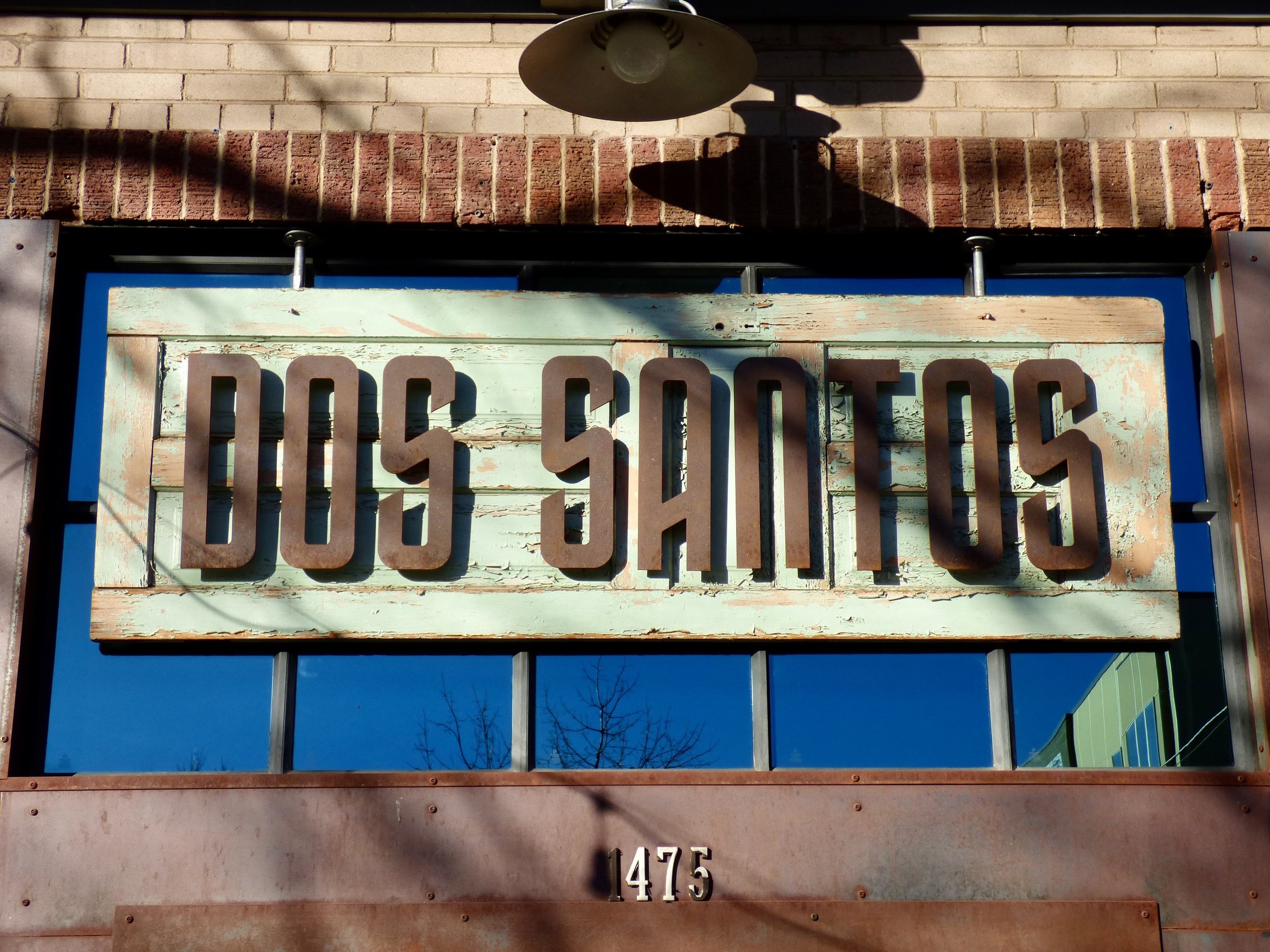 Sign reading 'DOS SANTOS' mounted on a brick building exterior with shadows cast on it and a blue sky reflected in a window below at Dos Santos, Denver, CO.