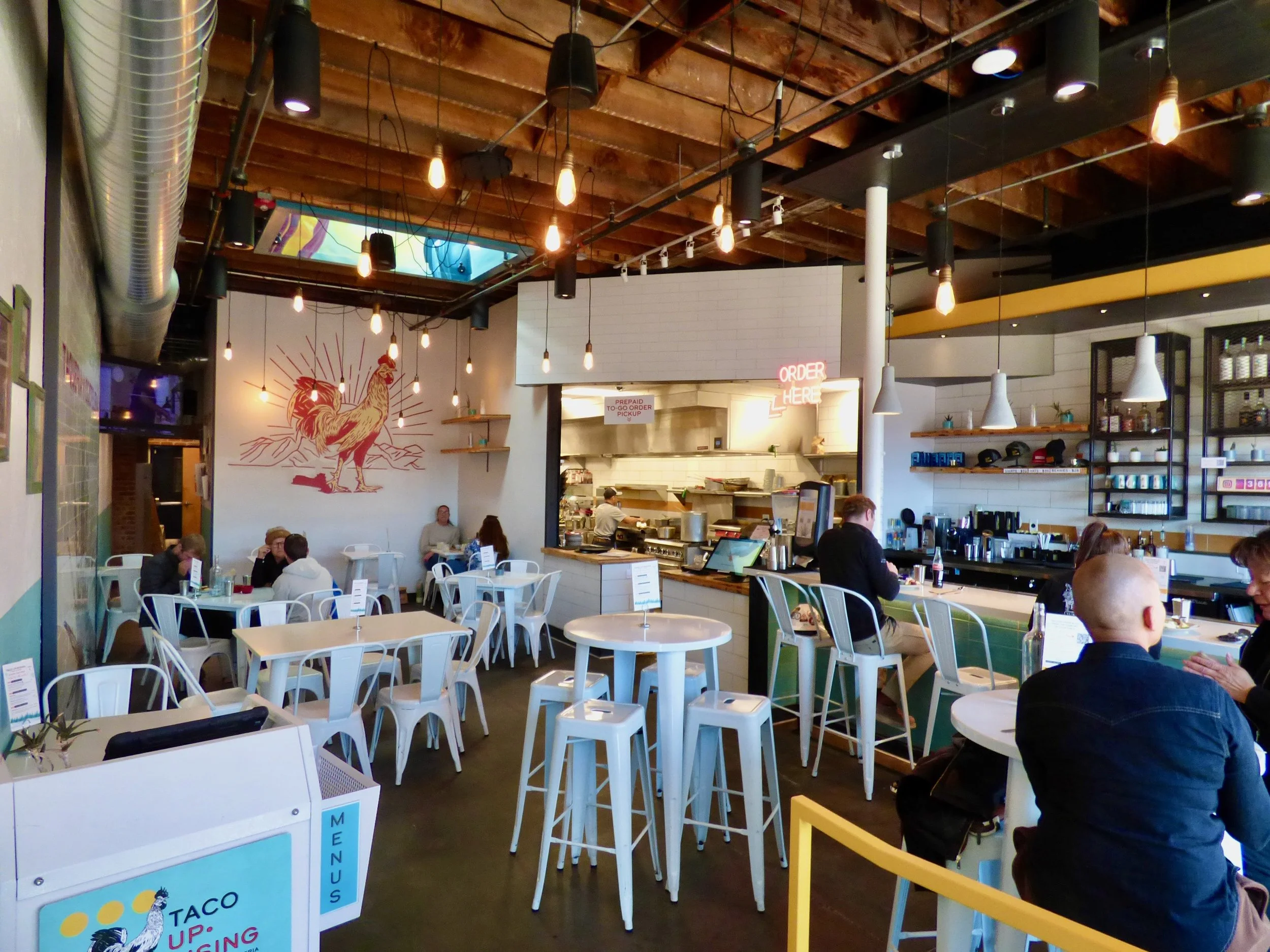 Interior of a modern restaurant with white tables and chairs, a bar counter, and various customers dining. A large mural of a rooster on the wall and warm pendant lighting hanging from the wooden ceiling.