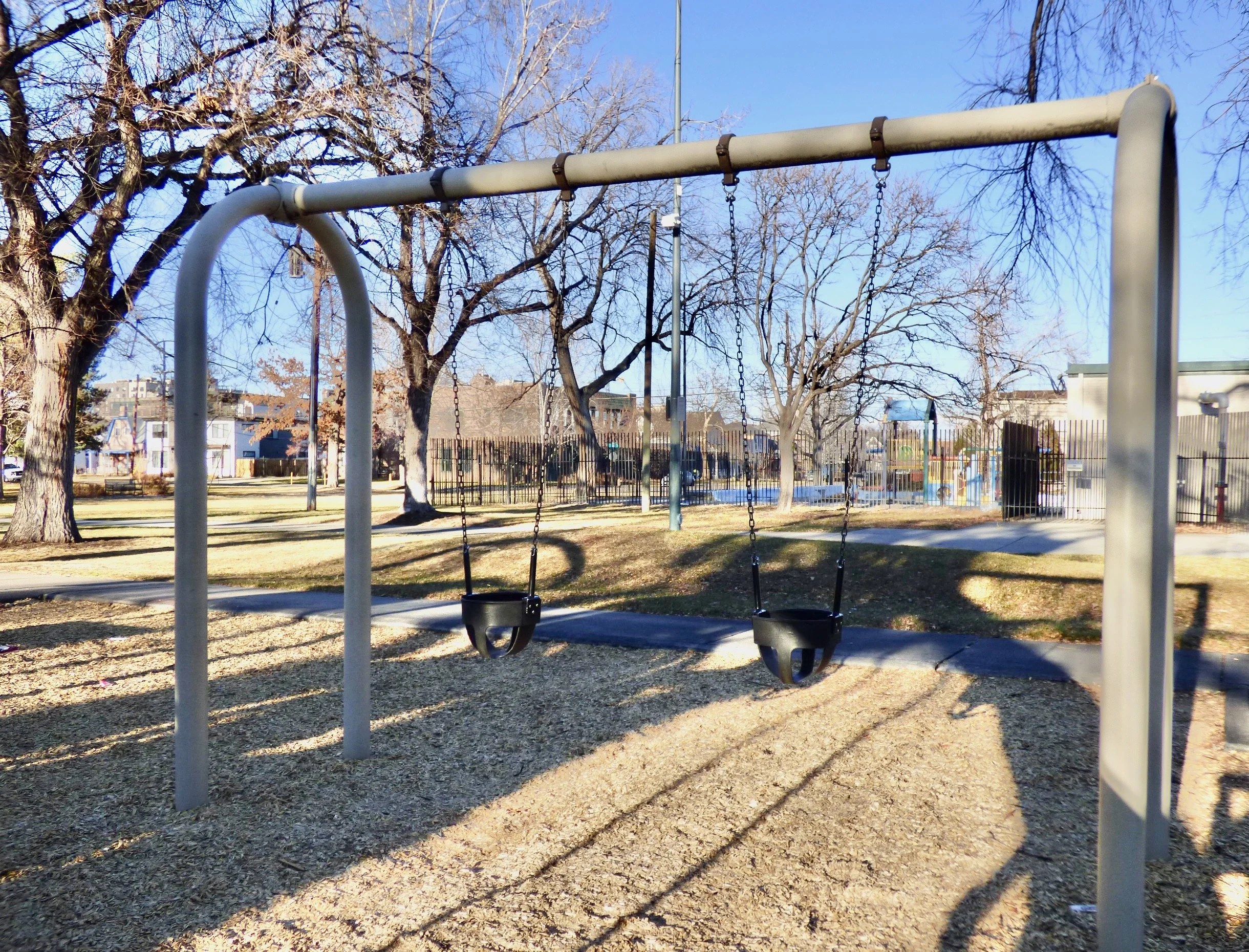 Empty playground with swings, leafless trees, fences, and a clear blue sky.
