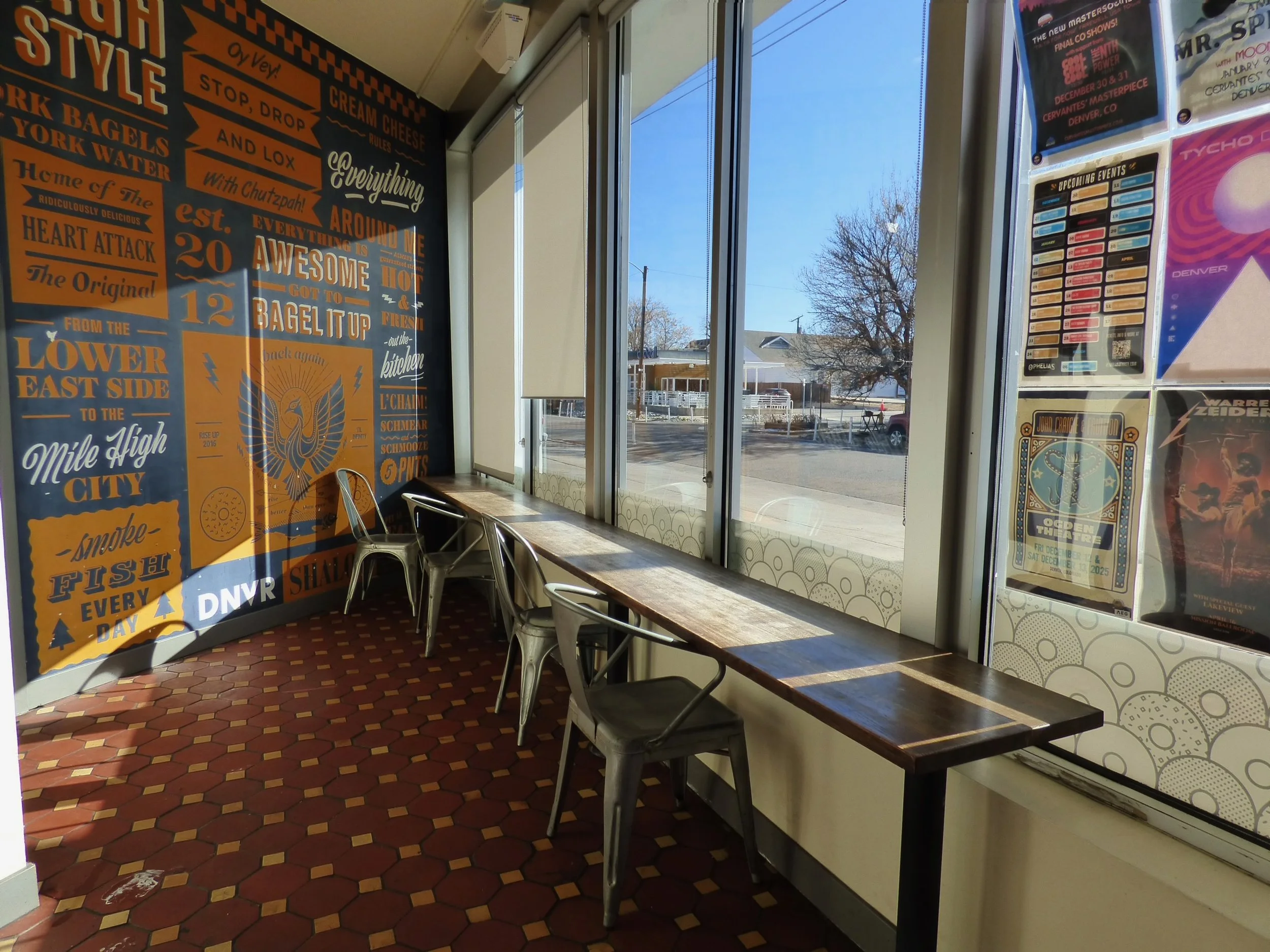 Interior of a café or restaurant with a large orange and black mural on the left wall featuring various phrases and designs, a long wooden counter next to large windows letting in sunlight, and a view of the street outside with houses and bare trees.