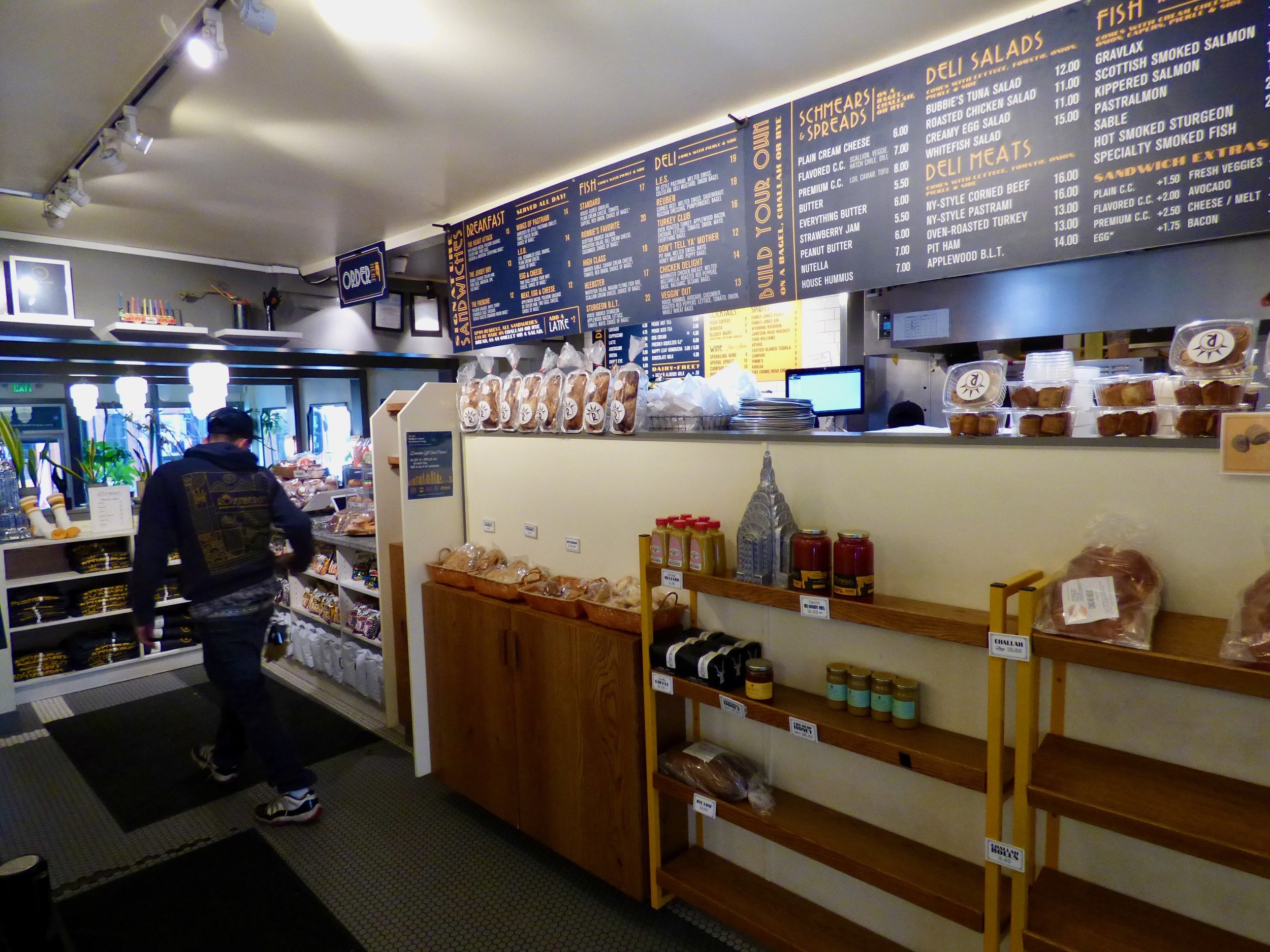 Interior of a cafe or deli with a menu board, shelves of packaged baked goods, and condiments on display. A customer is walking toward the counter.