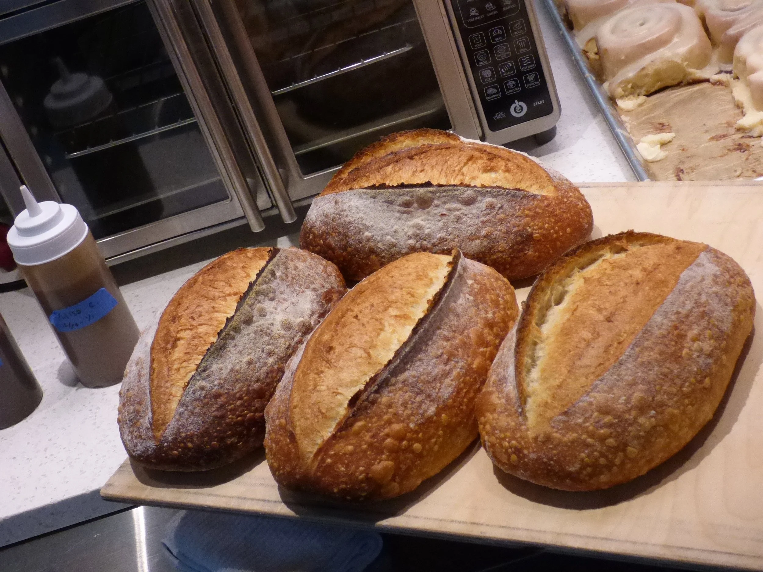 Four loaves of freshly baked bread with golden crust on a wooden cutting board, in a kitchen setting with appliances in the background at Elemental Bakery & Coffeehouse, Denver, CO 80205.