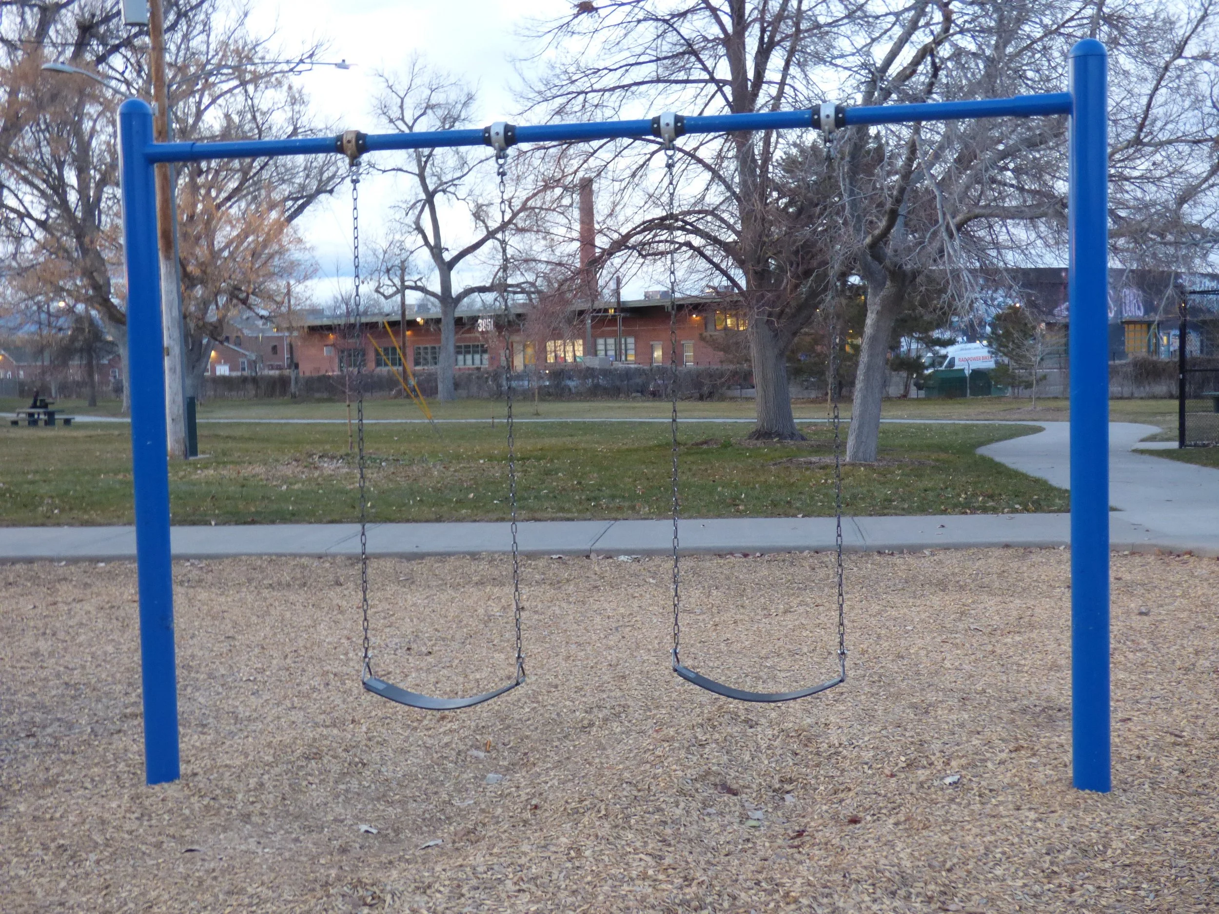 A playground with empty blue swings on a gravel surface, trees without leaves, a pathway, a grassy area, and residential buildings in the background in Martin J. Schafer Park, Denver, CO 80205.